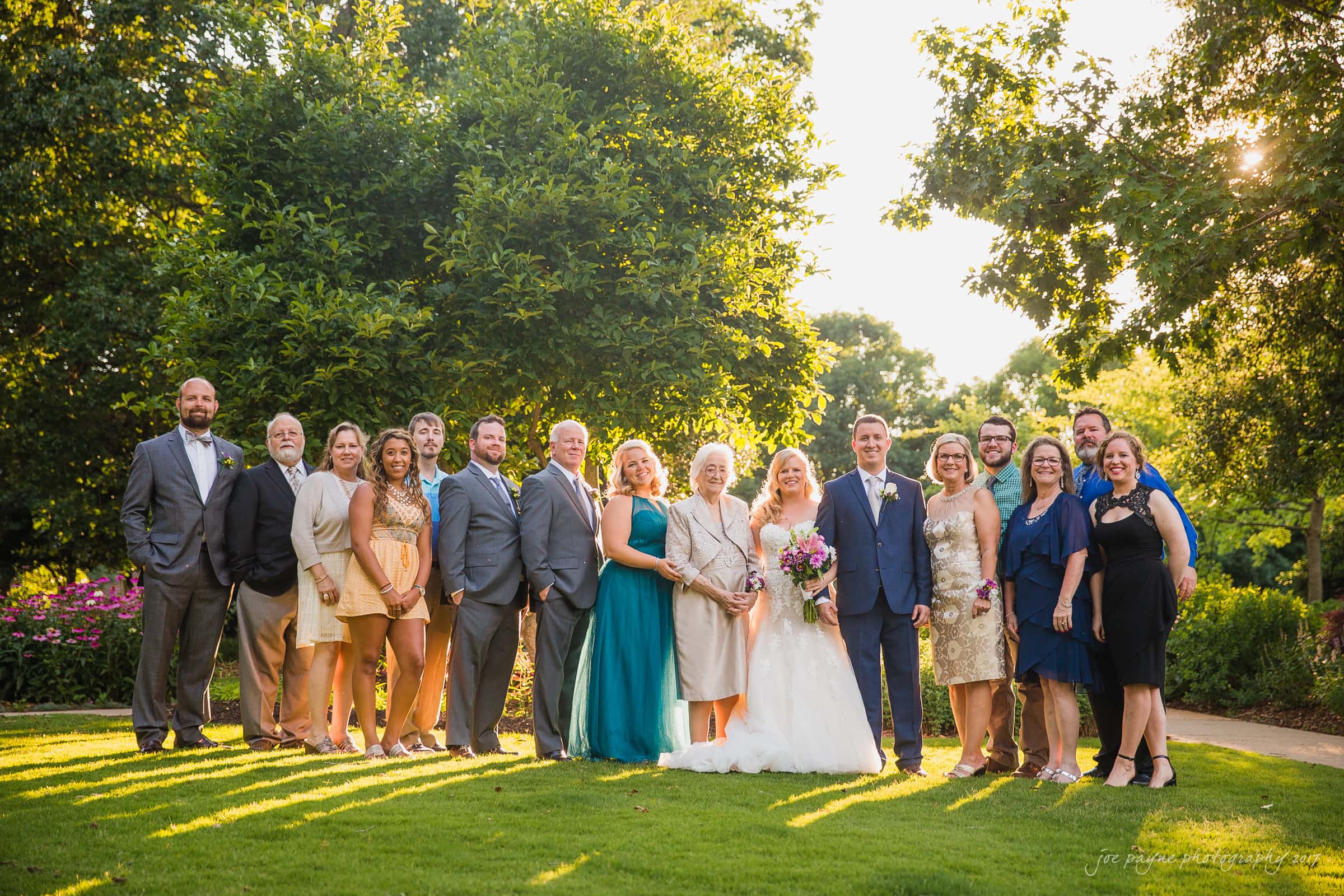 A group of people stand together in a garden setting, dressed formally. The bride and groom are in the center, with the bride holding a bouquet. The sun casts a warm glow on the scene, which is surrounded by trees and greenery.
