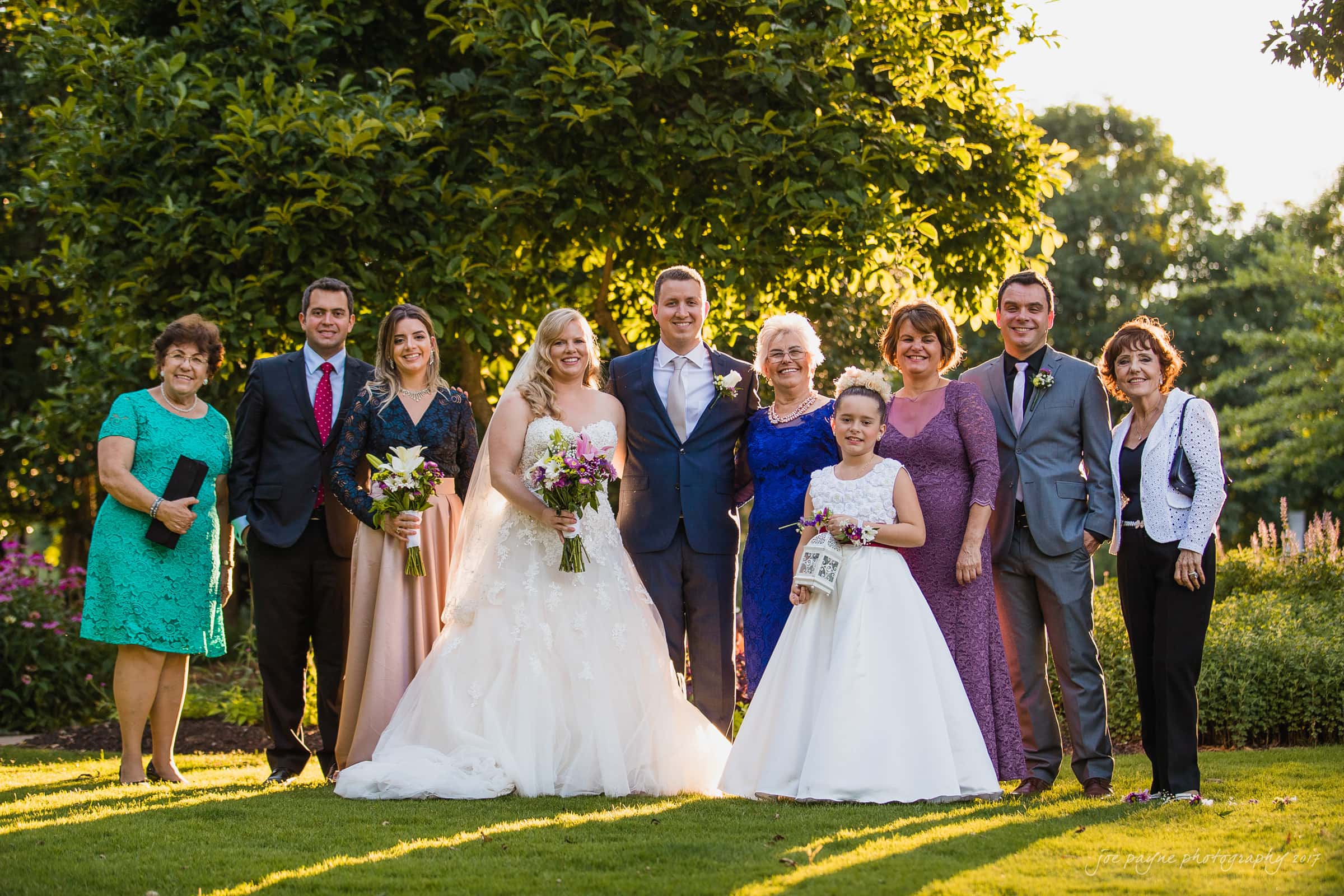 A wedding group photo outdoors with a bride in a white gown and a groom in a blue suit, surrounded by family and friends dressed in formal attire. They are posing on grass with trees and sunshine in the background.