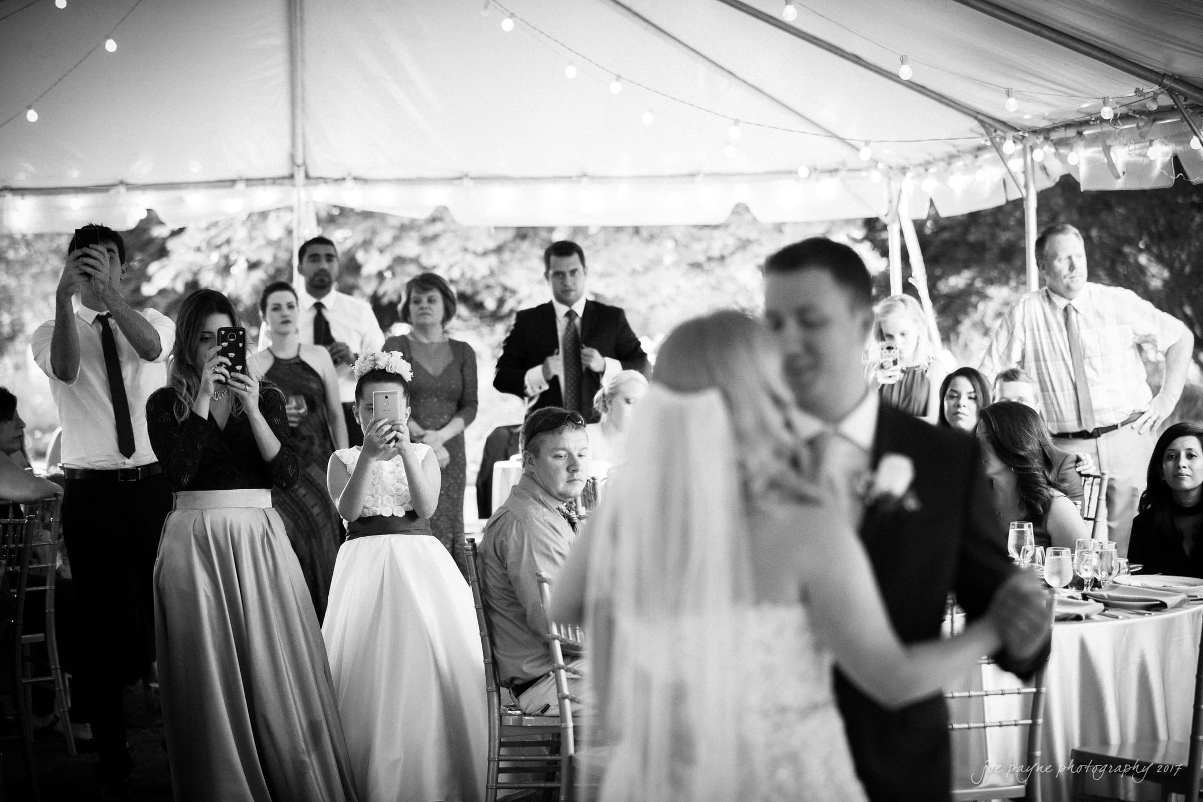 A bride and groom share a first dance under a tent, surrounded by seated guests and onlookers capturing the moment on their phones. The scene is in black and white, with string lights illuminating the background.