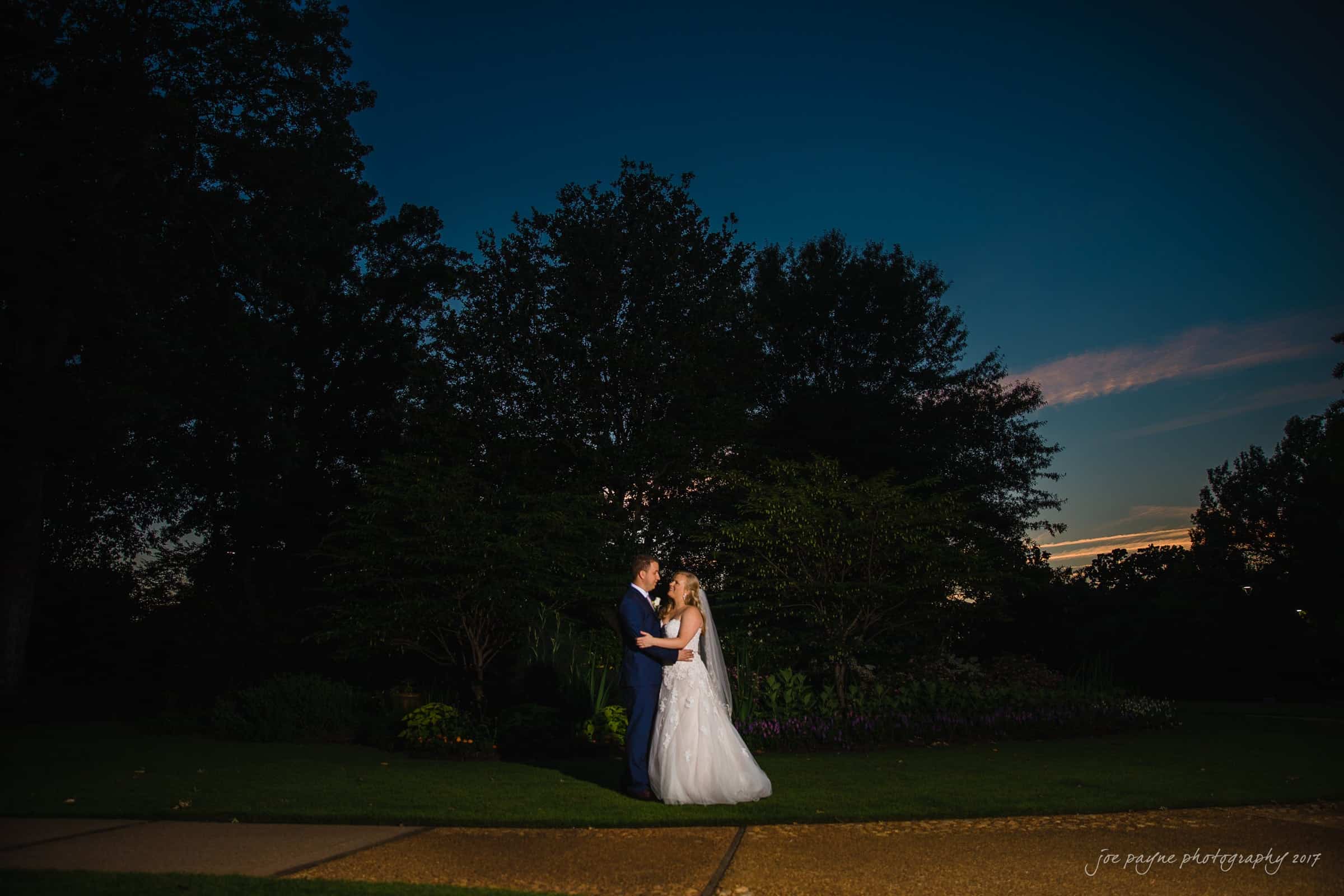 A bride and groom stand facing each other on a garden path during twilight. The bride is in a white dress, and the groom is in a dark suit. Trees and darkening sky form the background. Soft lighting highlights the couple in the serene setting.
