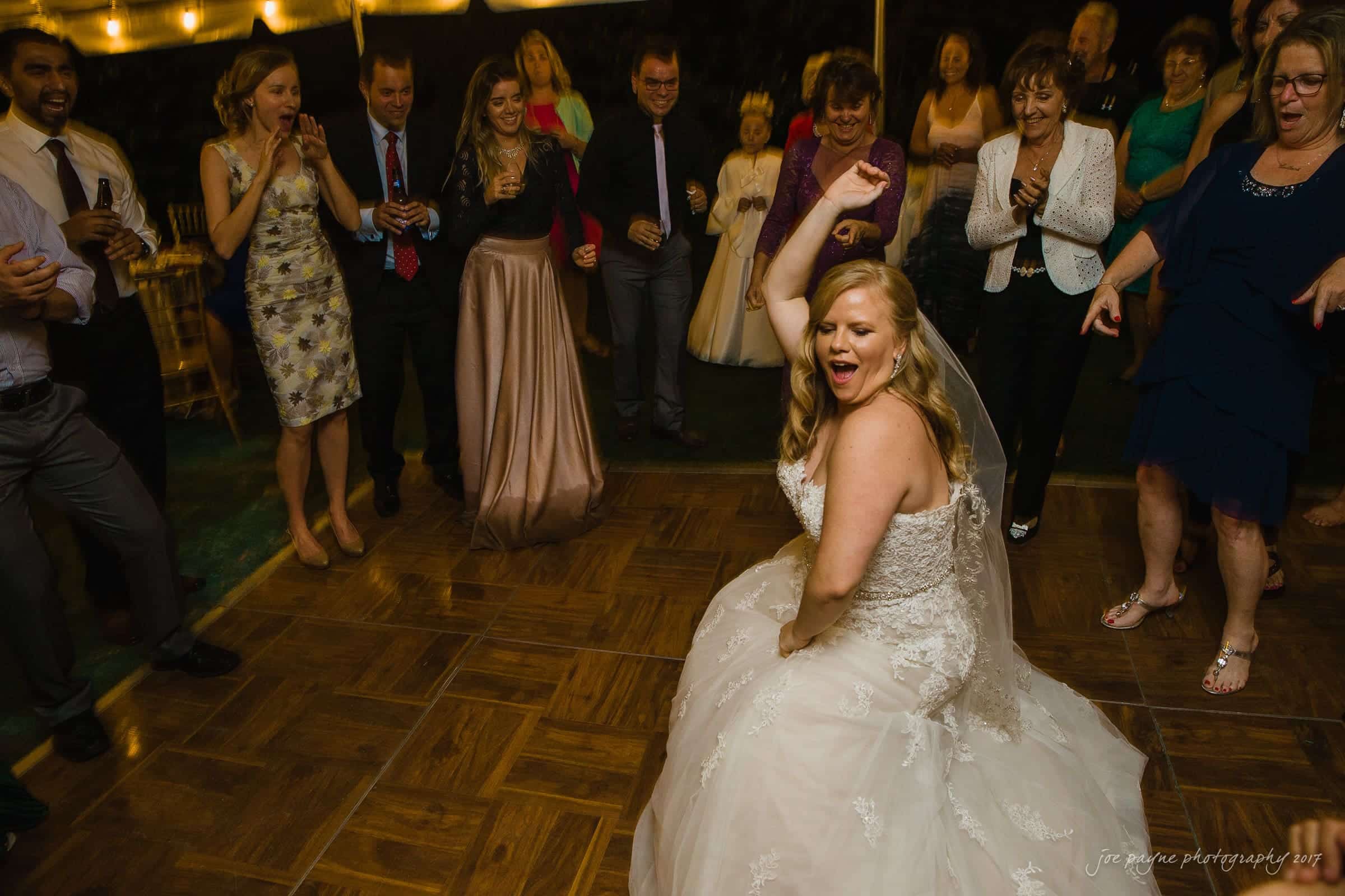 A bride in a white wedding dress enthusiastically dances on a wooden floor. Guests, dressed in formal attire, surround her, smiling and watching the celebration. String lights are visible in the background, adding a festive atmosphere.