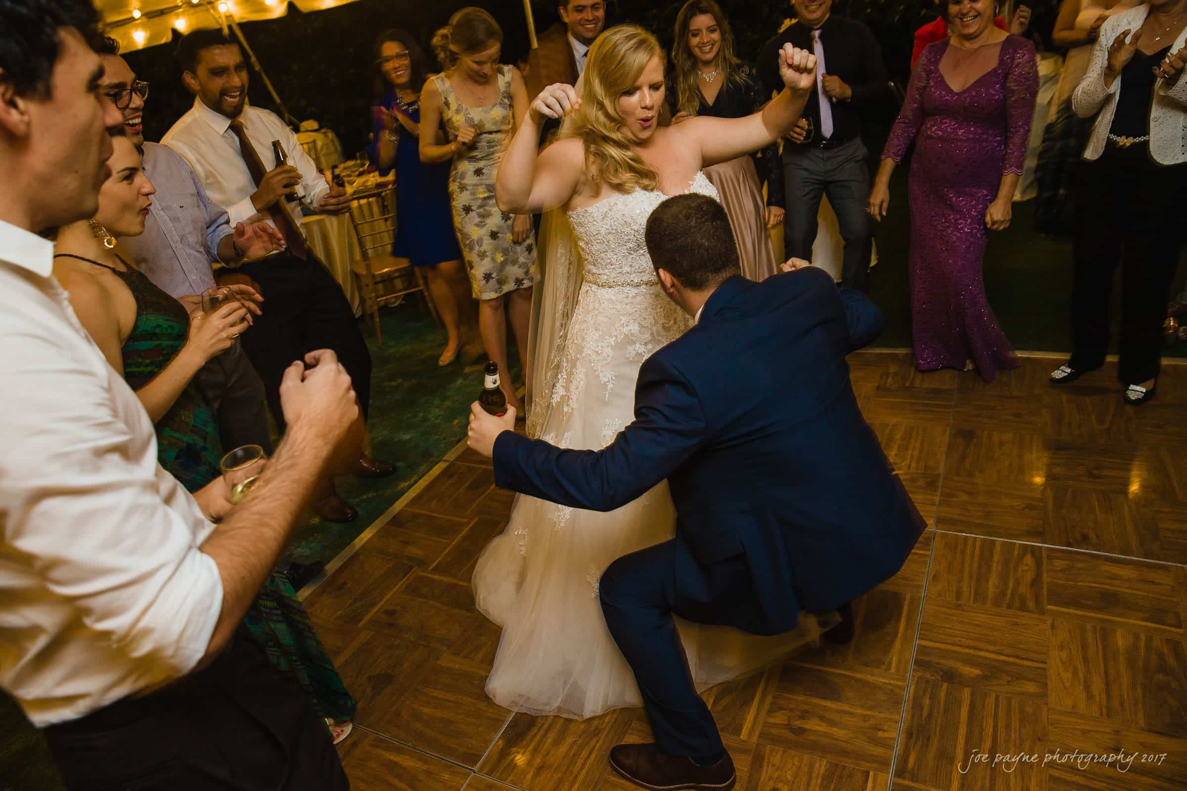 A joyful wedding reception scene with a bride in a white gown dancing energetically on a wooden floor. A man in a blue suit dances playfully in front of her. Guests surround them, clapping and smiling, under warm string lights.