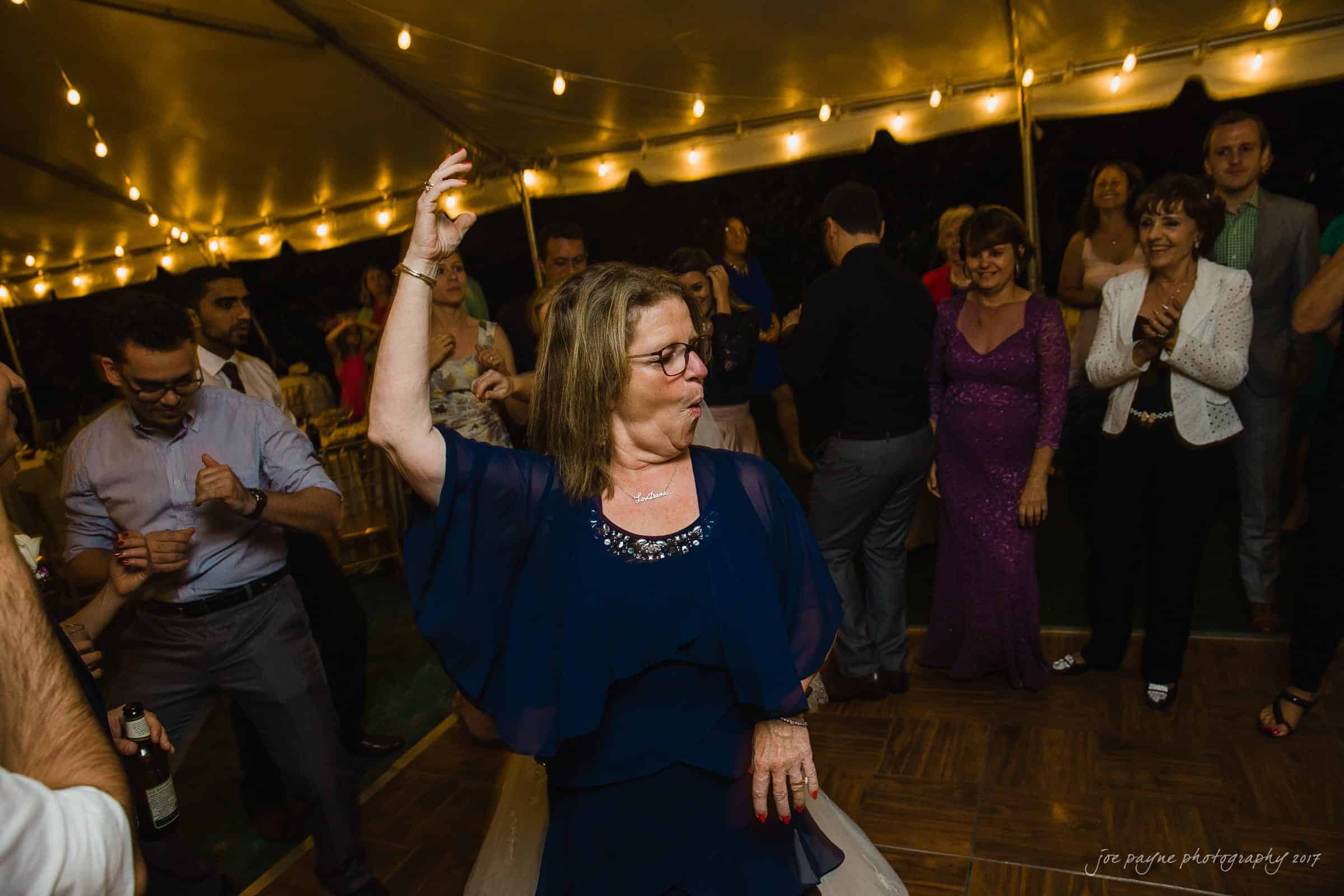 A woman in a navy dress dances energetically at a party under a tent adorned with string lights. People in formal attire surround her, enjoying the festive atmosphere.