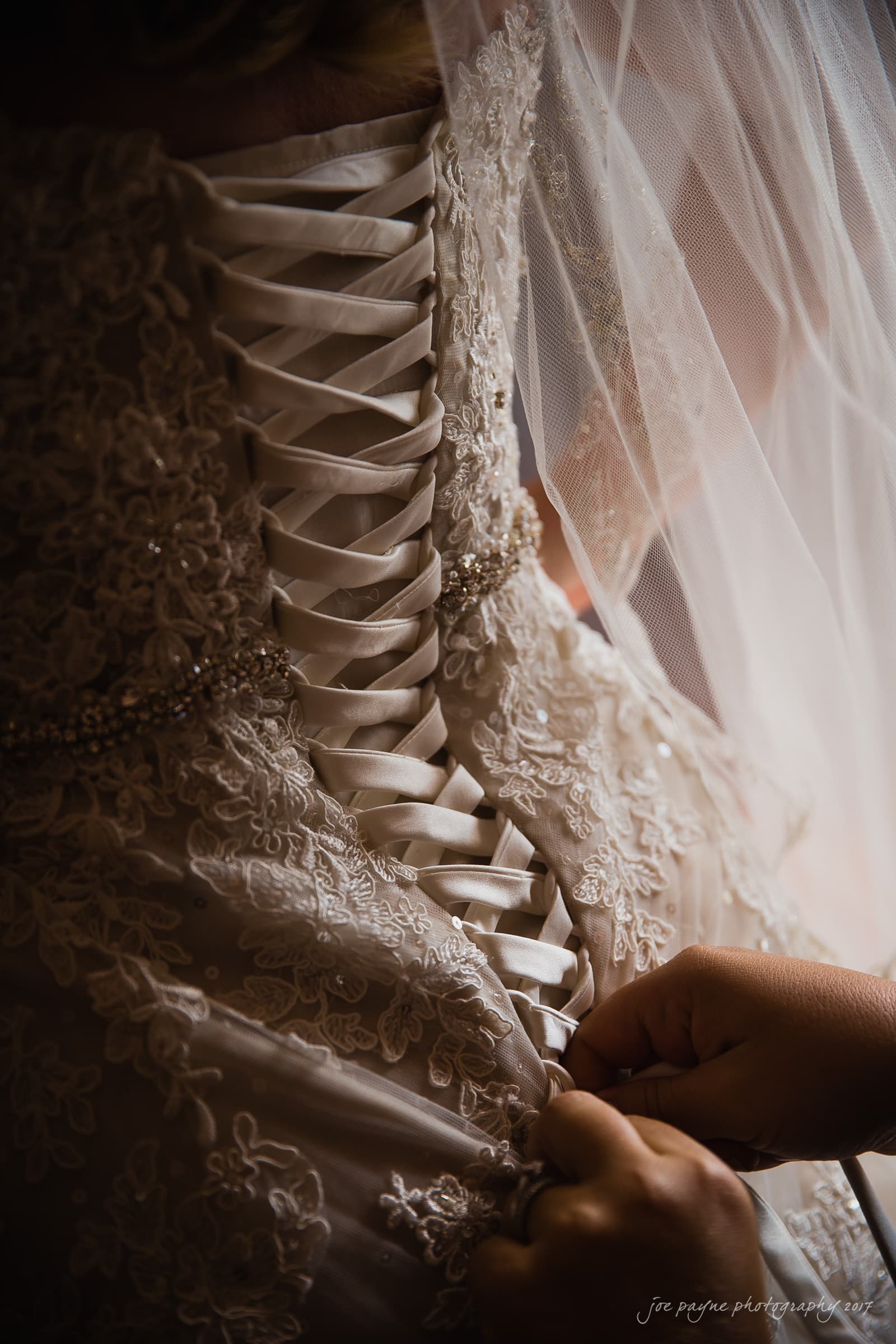 A close-up of a brides lace-up wedding dress being adjusted. The back of the dress features intricate lace details and satin ribbons. A veil is partially visible, and hands are tightening the laces.