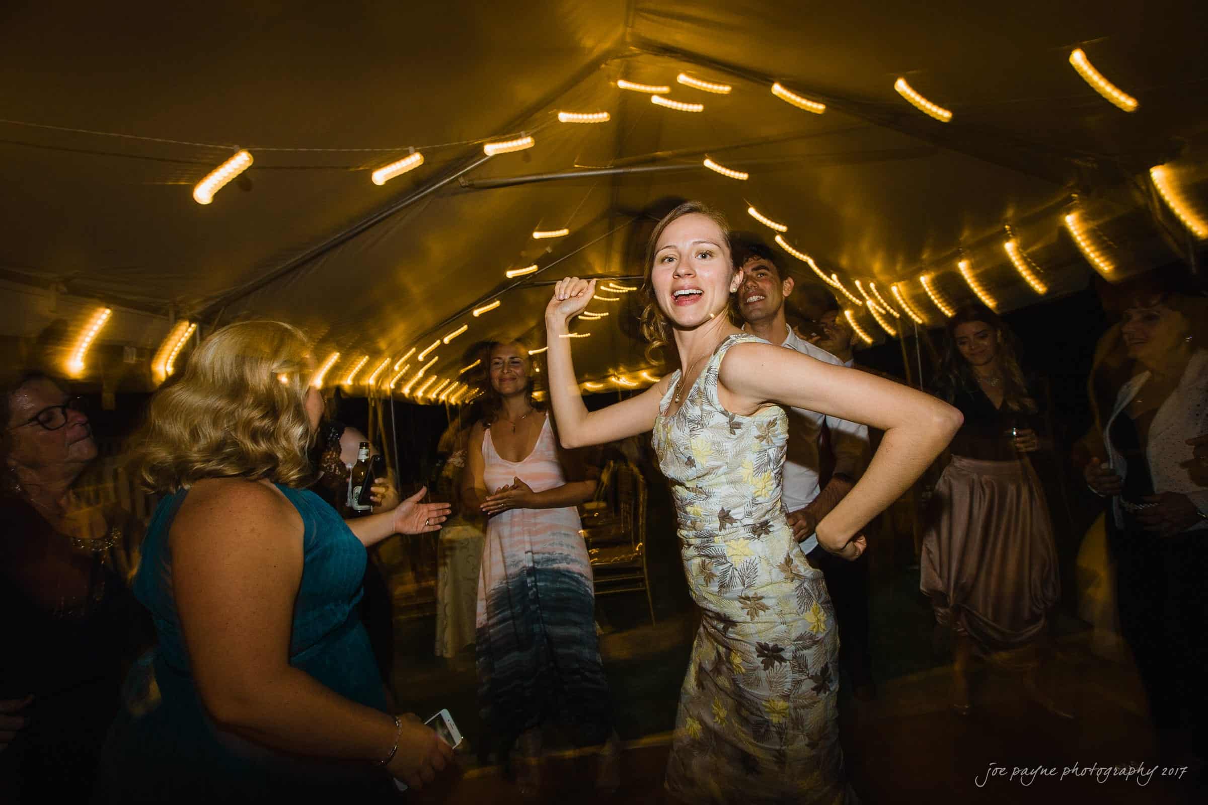 A group of people dancing under a tent with string lights. A woman in a patterned dress is at the center, smiling and raising her arm. The atmosphere is festive and the lighting creates a warm ambiance.