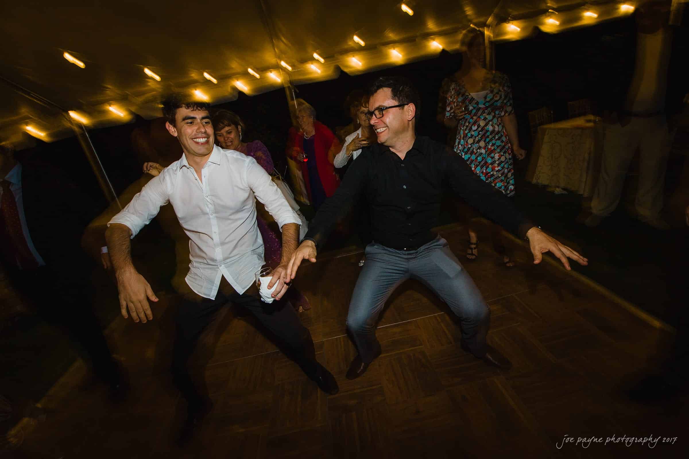 Two men are joyfully dancing at an indoor event. They are surrounded by other people watching. Strings of warm lights illuminate the venue, creating a festive atmosphere.