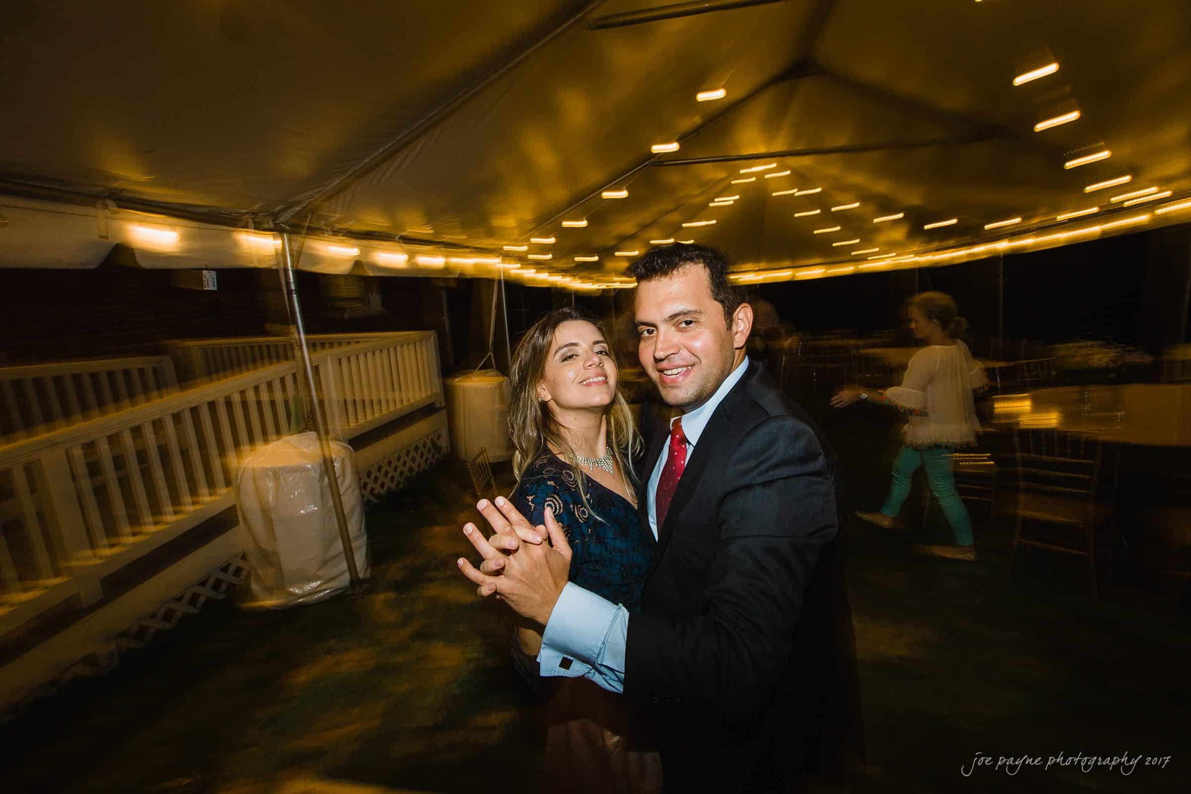 A couple smiling and dancing under a tent with string lights. The man is wearing a suit and red tie, while the woman is in a dark dress. The setting seems to be an evening event, with a walkway and tables in the background.