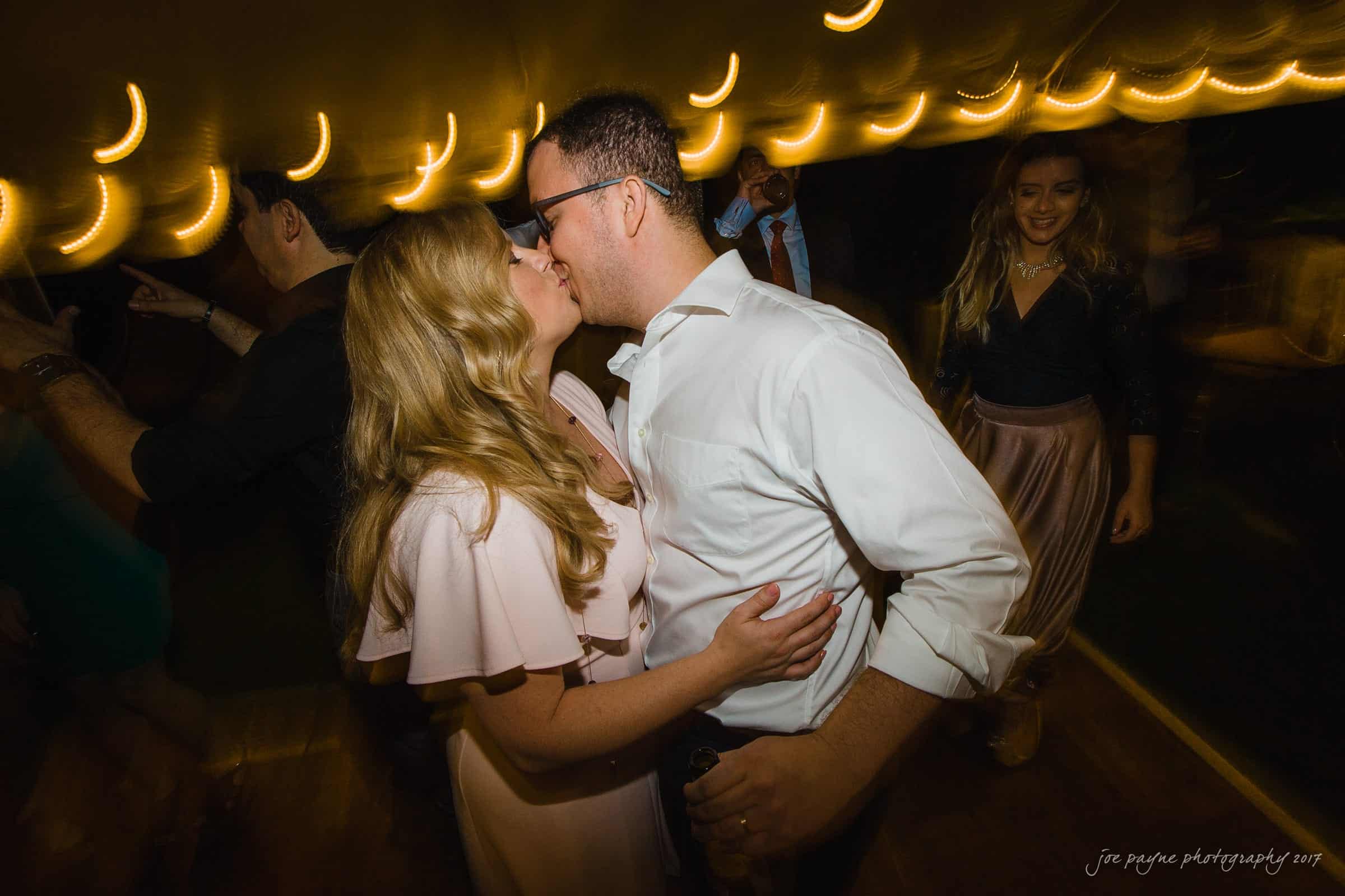 A couple shares a kiss on a dimly lit dance floor, surrounded by blurred figures and string lights, creating a romantic atmosphere.