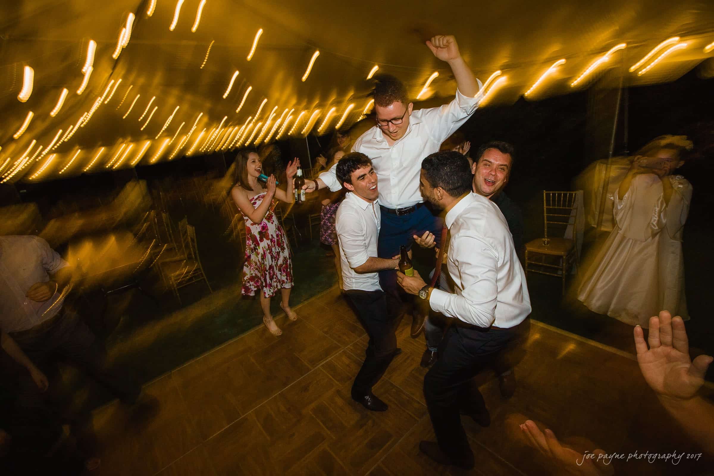 A lively party scene with guests dancing energetically under string lights. One person is lifted into the air by others, all smiling and celebrating. The atmosphere is joyful and festive.