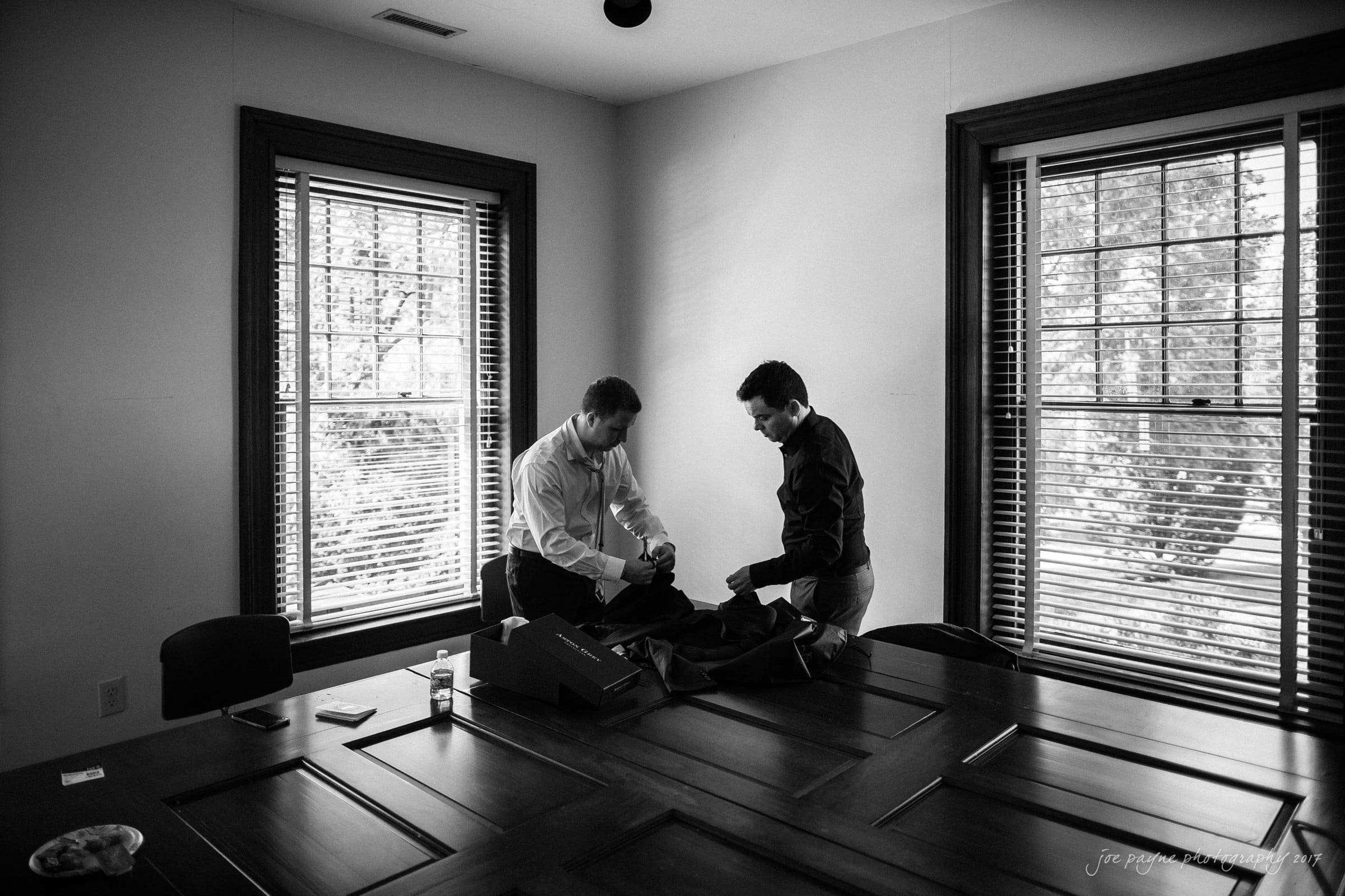 Two men stand at a wooden table in a room with large windows. They are engaged in a task, possibly assembling or packing something. The room is well-lit with natural light streaming through the blinds.