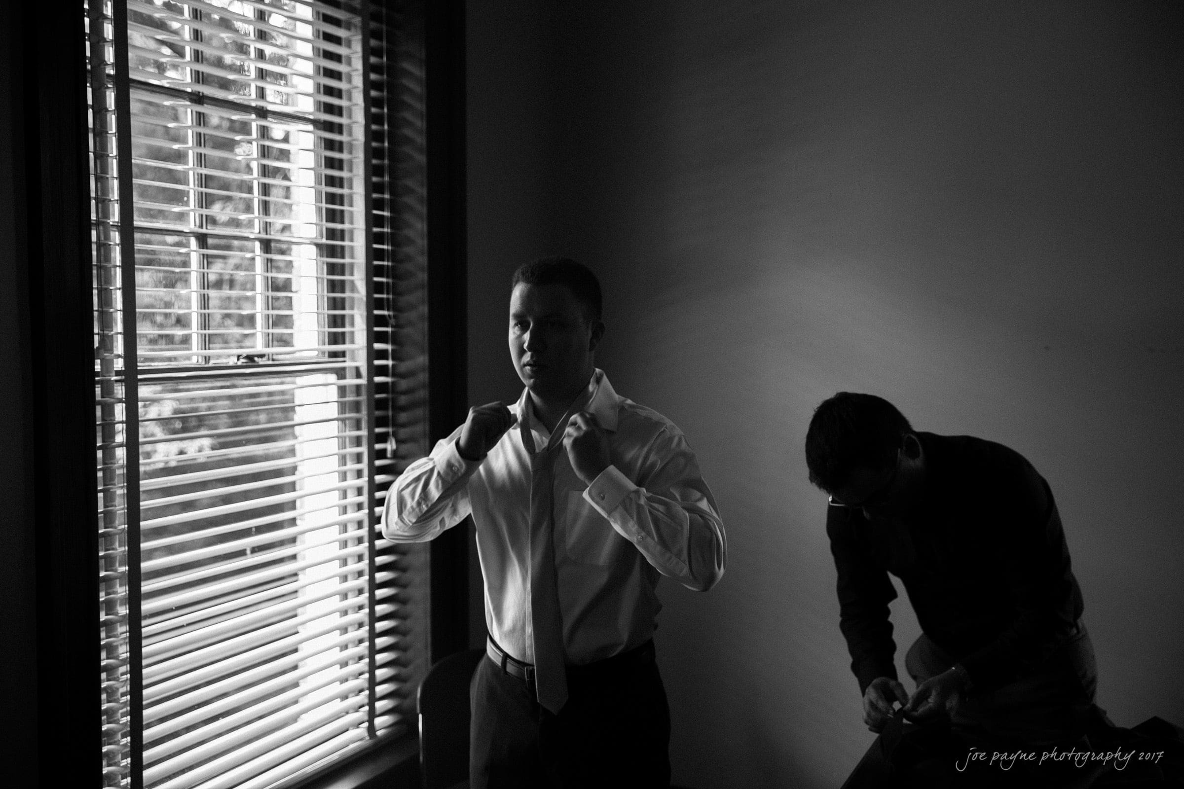 A man adjusts his tie in a dimly lit room near a window with blinds, casting striped shadows. Another person stands in the background, looking down and holding an object. The scene is captured in black and white.