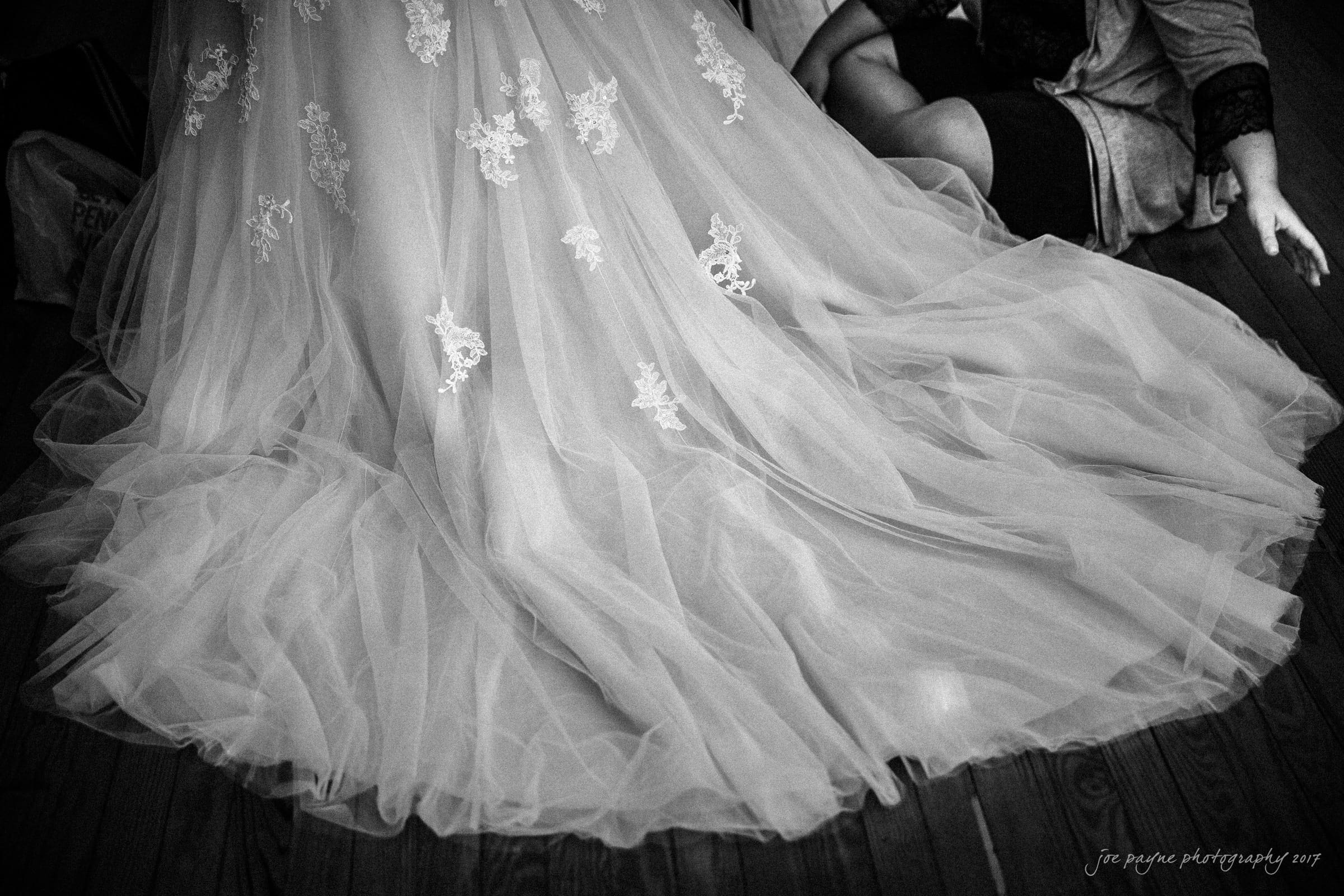 A black and white image of a person in a wedding dress with lace details, seated on a wooden floor. The flowing gown spreads elegantly around them, and another individual is partially visible next to them.