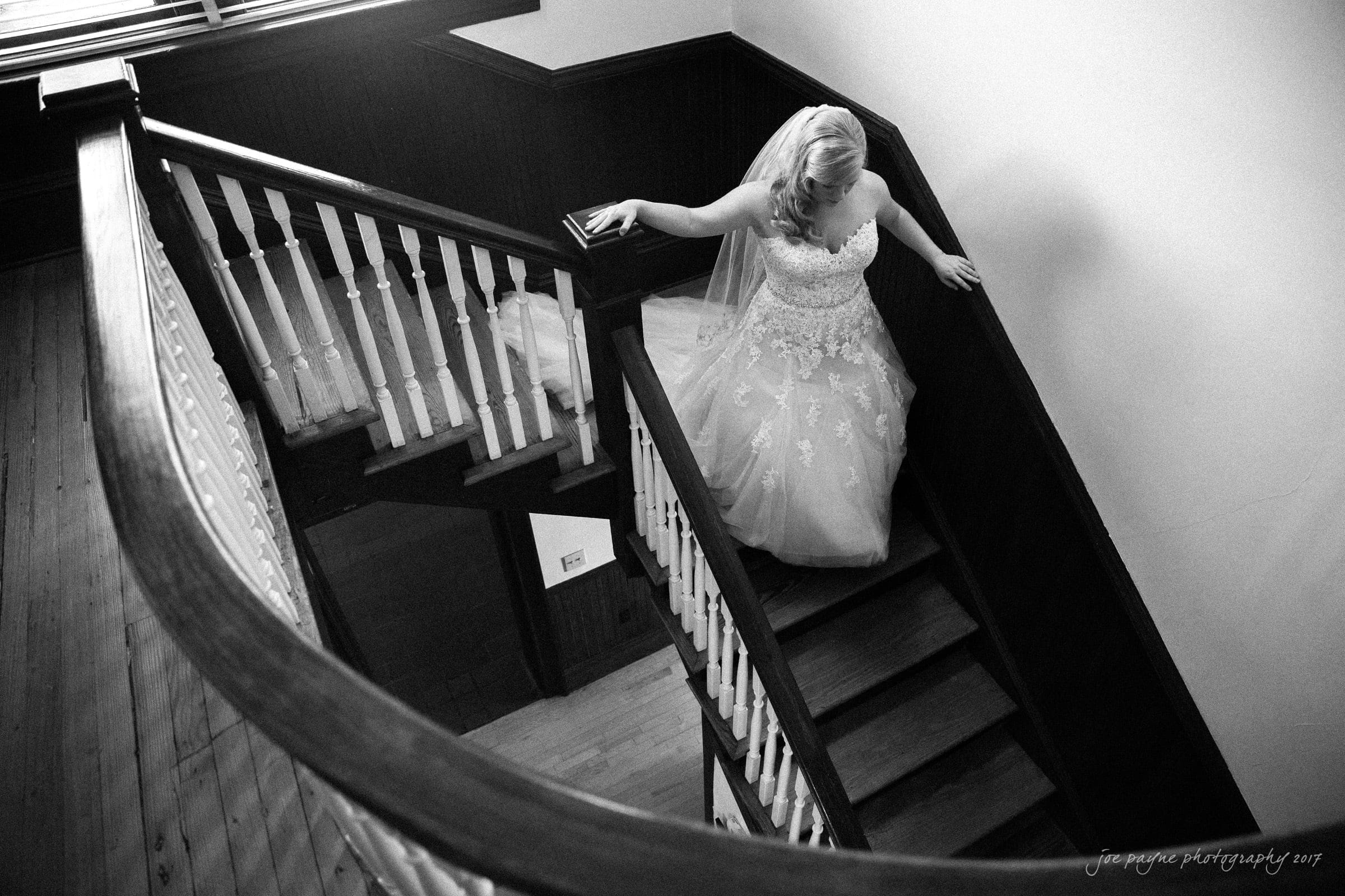 A bride in a lace wedding gown descends a wooden staircase. Her hand gently touches the railing. The scene is captured in black and white, highlighting the intricate details of her dress and the classic style of the staircase.
