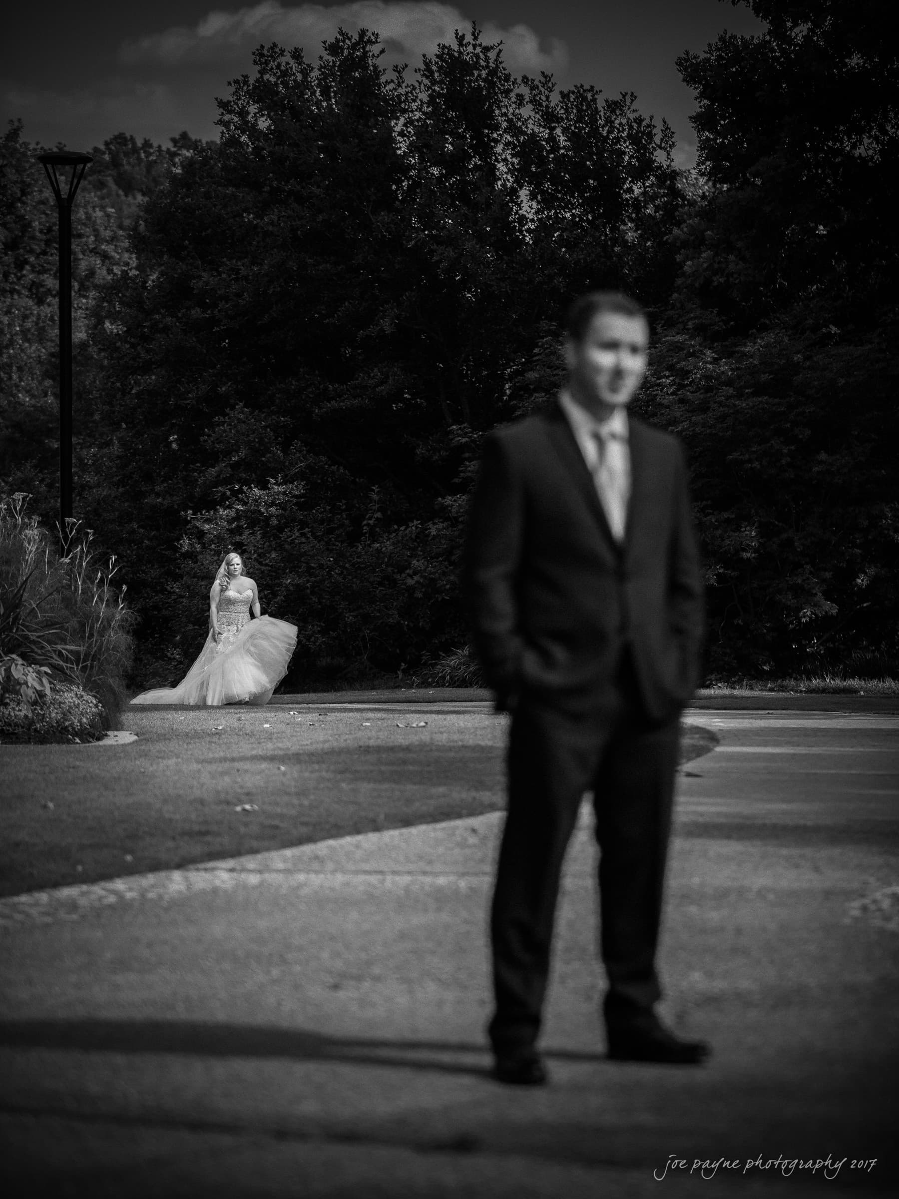 A groom in focus stands in the foreground, while a bride walks towards him in the background on a tree-lined path. The scene is in black and white, creating a timeless and romantic atmosphere.