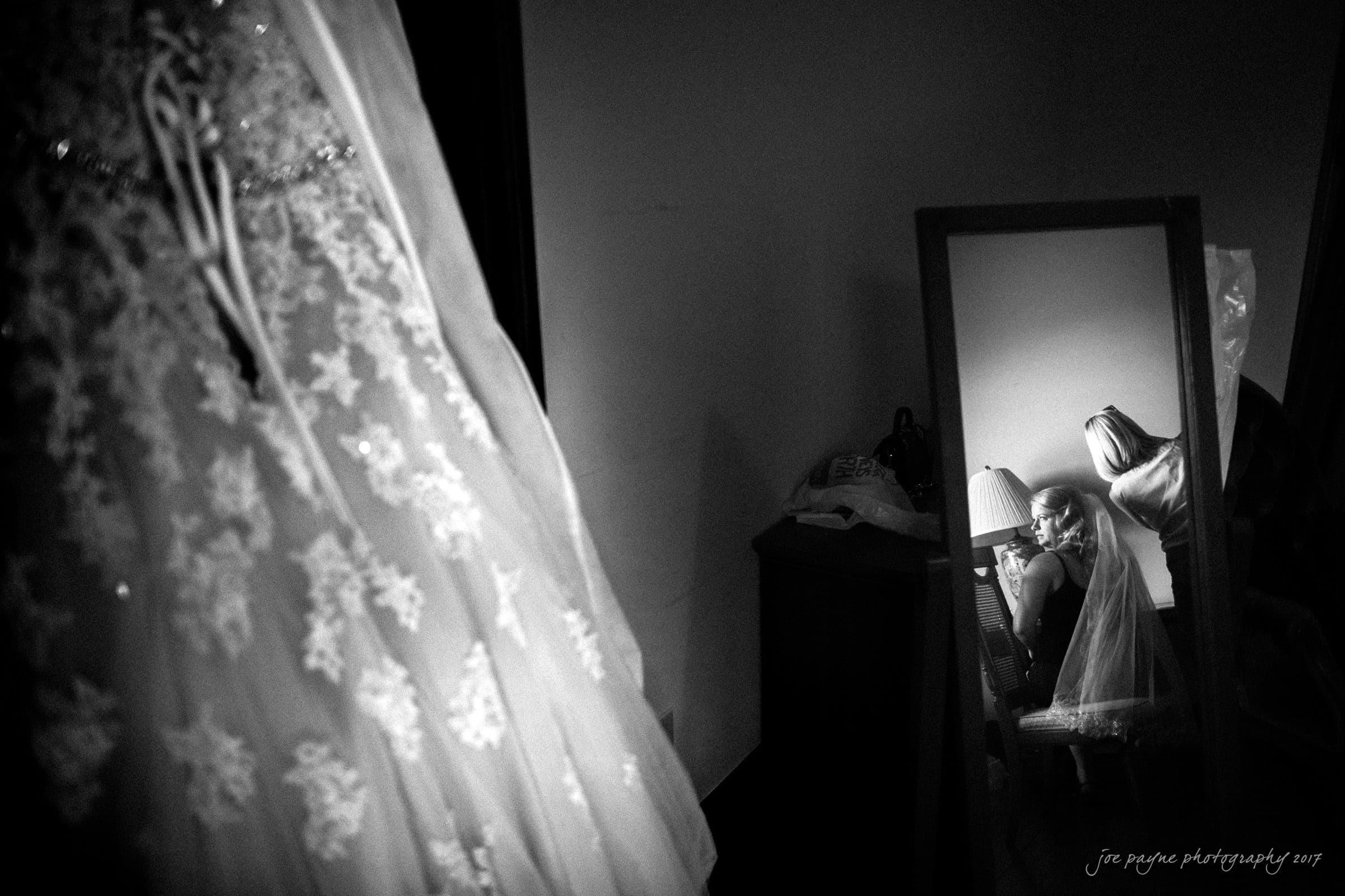 Black and white photo of a bride wearing a veil, sitting at a vanity mirror. The foreground shows an out-of-focus wedding dress. The room is softly lit, creating a serene and intimate atmosphere.