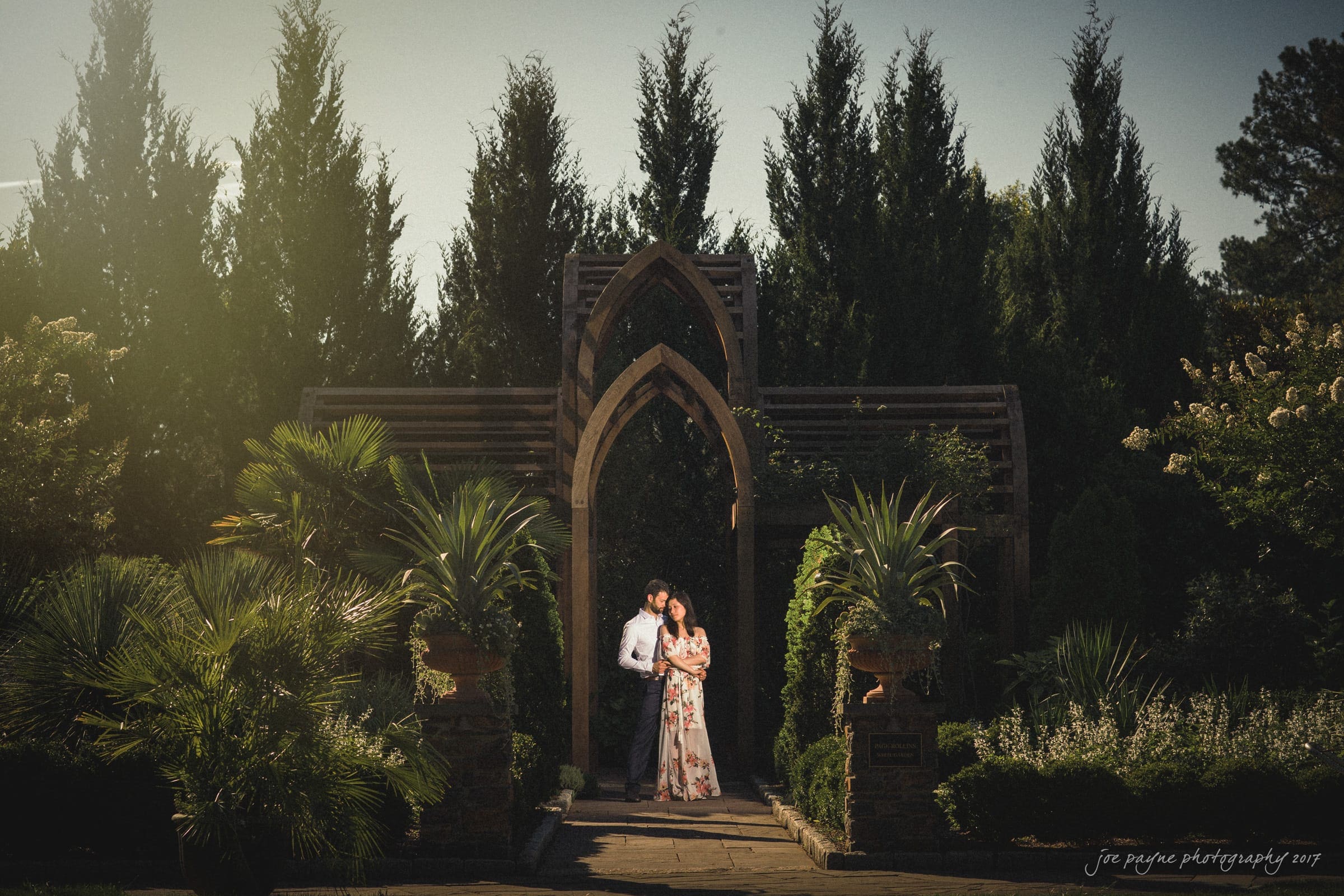 A couple stands embraced under an ornate wooden archway in a lush garden. Sunlight filters through tall trees, illuminating the scene. The woman wears a floral dress, and the man is in a white shirt and tie. The atmosphere is serene and romantic.
