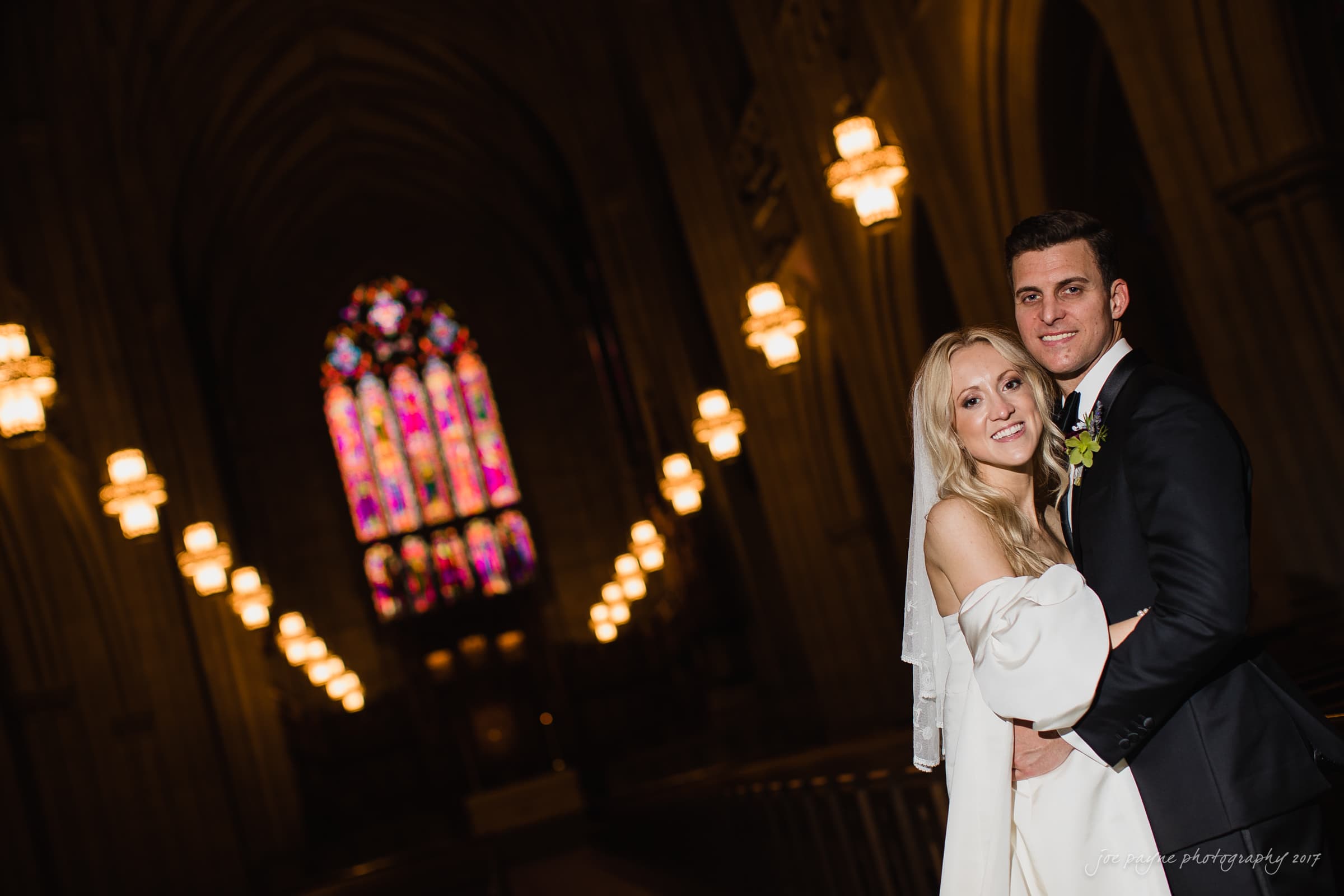 Duke Chapel Weddings A professionally lit wedding portrait inside Duke Chapel, using flash to illuminate the happy couple while darkening the interior to showcase the vibrant colours of the stained-glass window and glowing pendant lights.