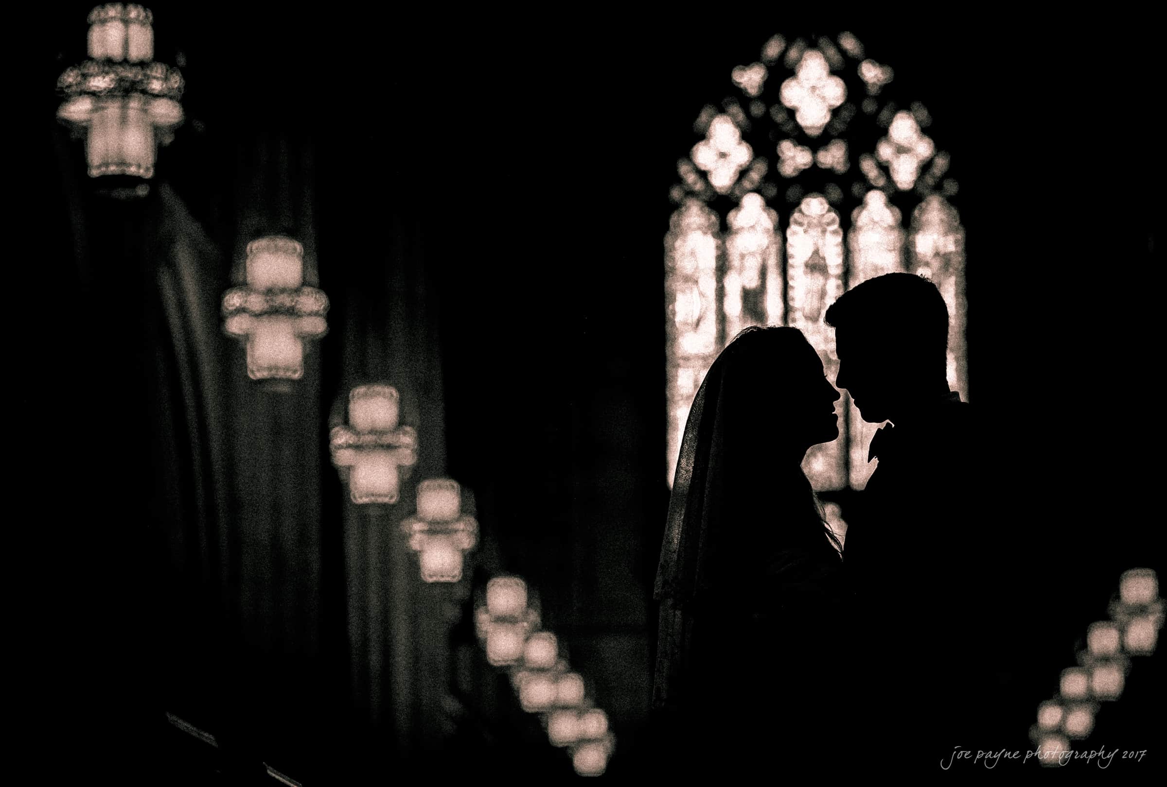 Silhouetted couple facing each other in a dimly lit church, with illuminated chandeliers and a large stained glass window in the background. The scene evokes a romantic, serene atmosphere.
