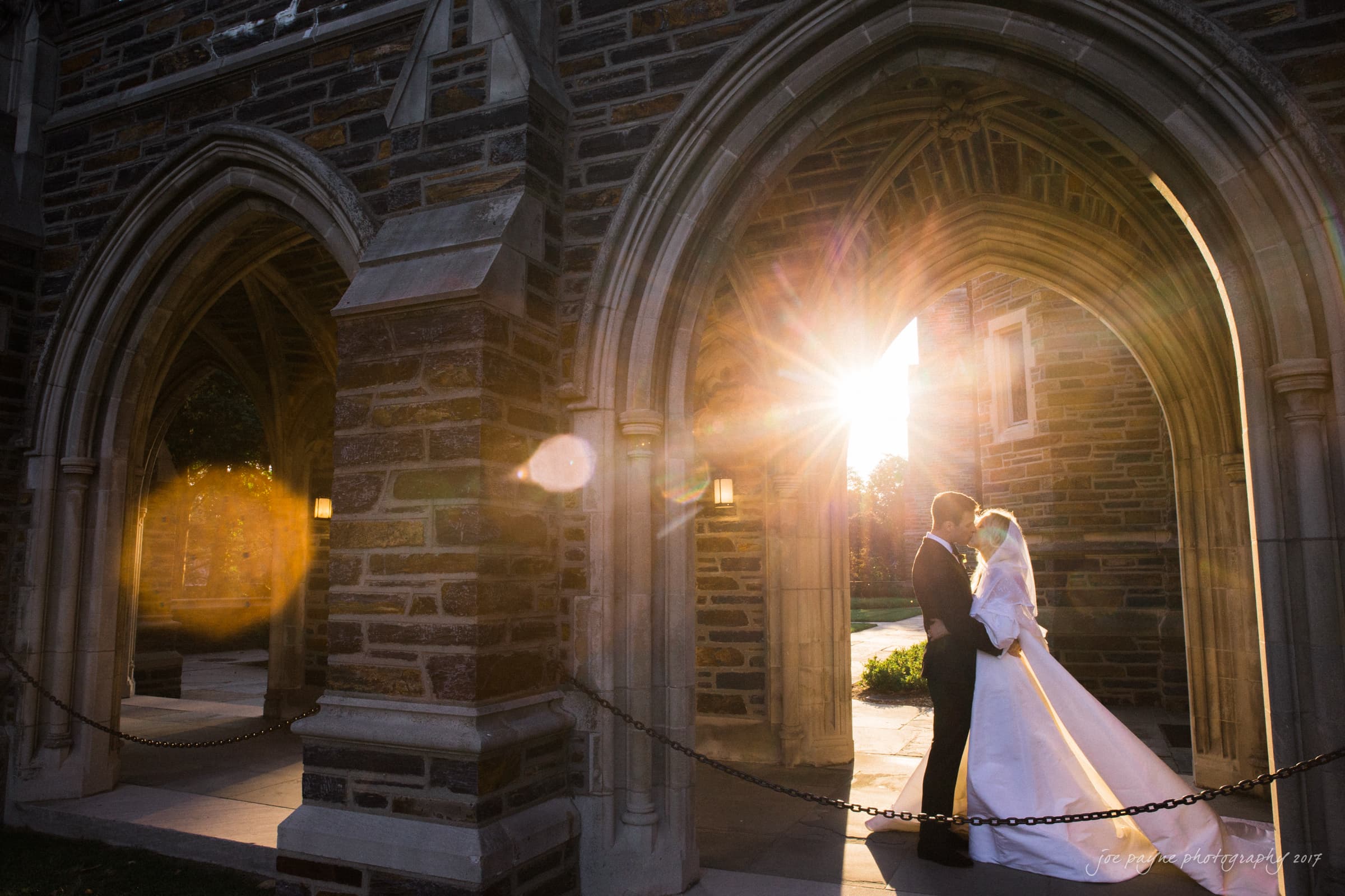 Duke Chapel Weddings Duke Chapel WeddingsA romantic sunset wedding portrait of Lara and Juice kissing in the Duke Chapel arcades, beautifully illuminated by a warm, backlit lens flare from the setting sun.