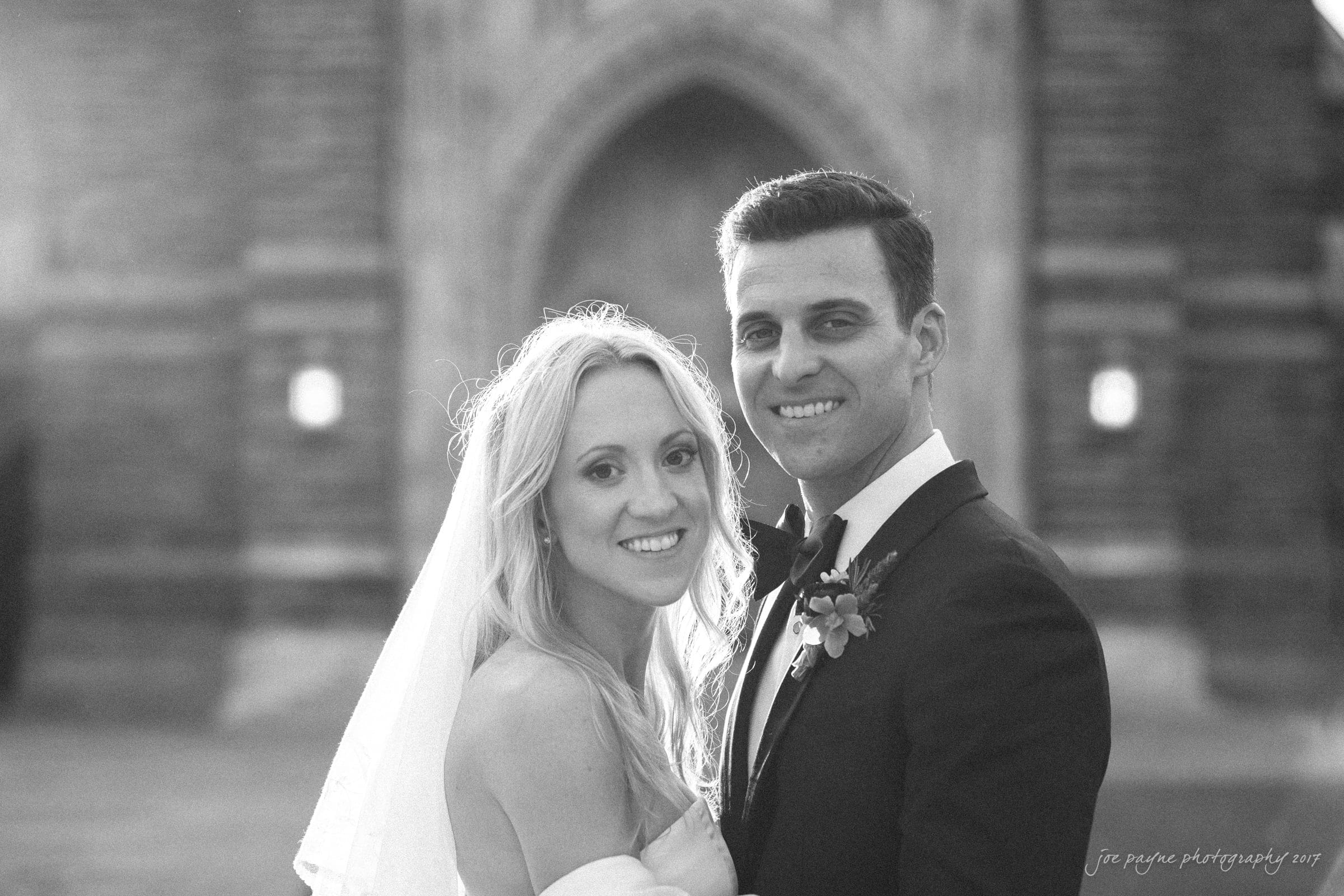 Duke Chapel Weddings A classic black and white wedding portrait of the couple smiling at the camera, with the grand stone façade and towering entrance of Duke Chapel rising majestically in the background.