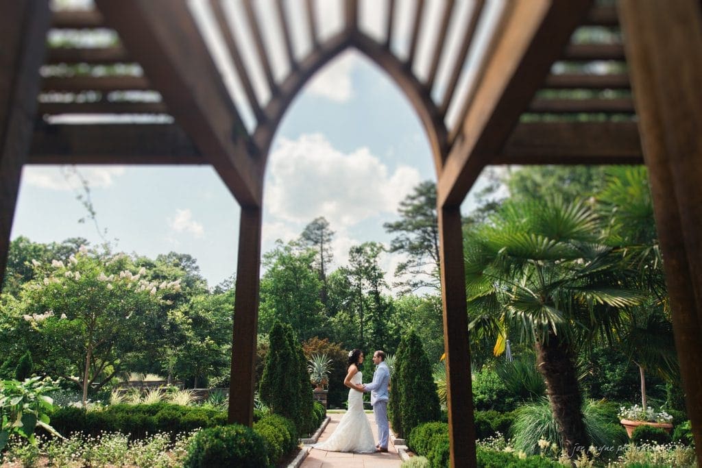 duke gardens wedding photography - jordan & zeke A couple embraces under a wooden archway in a lush garden. The pathway is lined with greenery and trees, with a bright, partly cloudy sky above. The scene evokes romance and tranquility.