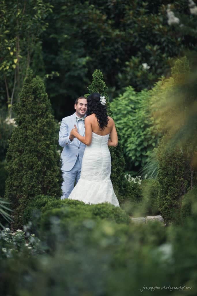 duke gardens wedding photography - jordan & zeke A bride in a white dress and a groom in a light blue suit hold hands, smiling at each other in a lush, green garden setting with tall trees and hedges surrounding them.