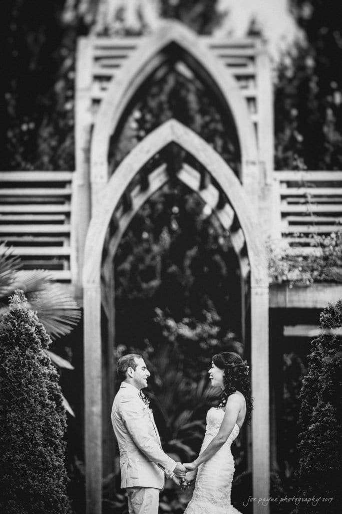duke gardens wedding photography - jordan & zeke A bride and groom stand holding hands outdoors under a decorative archway. The bride wears a long white dress, and the groom is in a suit. They face each other, surrounded by lush greenery. The photo is in black and white.