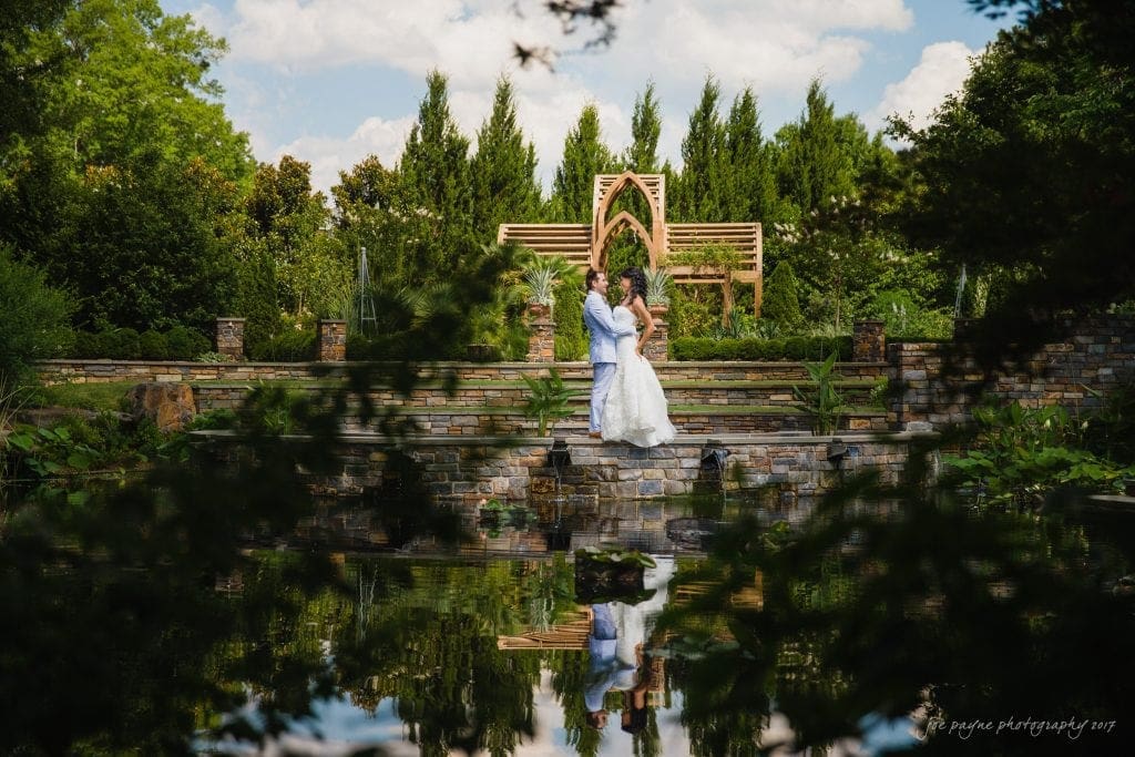 duke gardens wedding photography - jordan & zeke A couple in wedding attire shares a romantic moment on stone steps surrounded by lush greenery and a serene pond. In the background, a rustic wooden arch blends with tall trees under a partly cloudy sky.