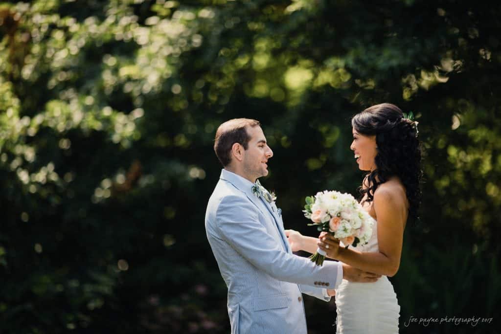 duke gardens wedding photography - jordan & zeke A bride and groom stand facing each other outdoors in a sunlit, wooded area. The bride holds a bouquet of white and pink flowers, and both are smiling warmly. The groom wears a light-colored suit.