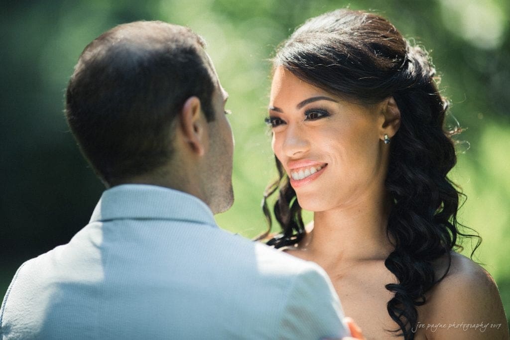 duke gardens wedding photography - jordan & zeke A couple stands closely together outdoors. The woman is smiling warmly at the man, who has his back to the camera. The background is lush and green, suggesting a natural setting.