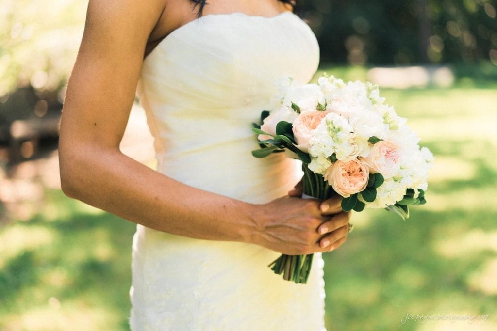 duke gardens wedding photography - jordan & zeke A bride in a strapless white wedding dress holds a bouquet of white and pale pink flowers. She is outdoors on a sunny day with green grass in the background.