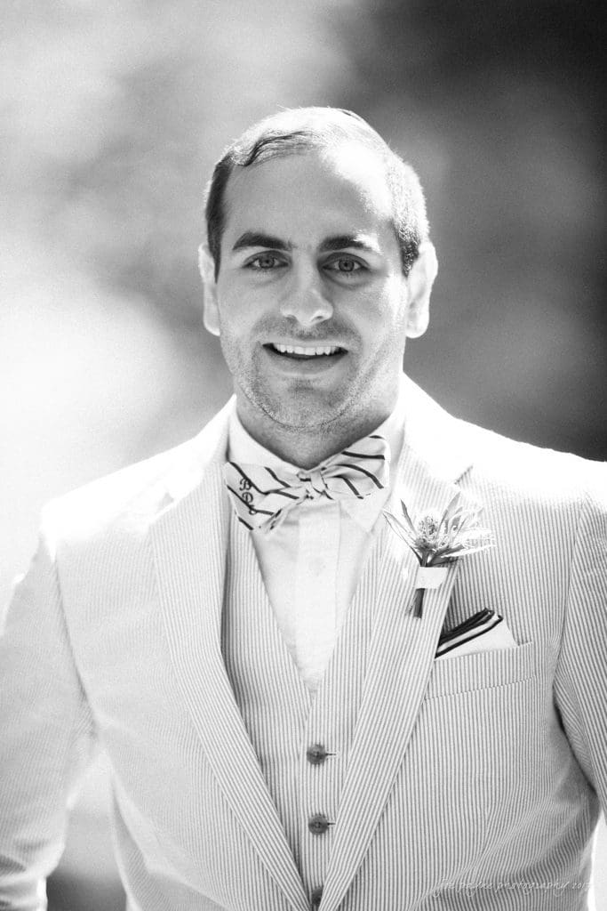 duke gardens wedding photography - jordan & zeke A man in a light-colored, striped suit with a bow tie smiles warmly. He wears a boutonniere on his lapel and stands against a softly blurred background. The image is in black and white.