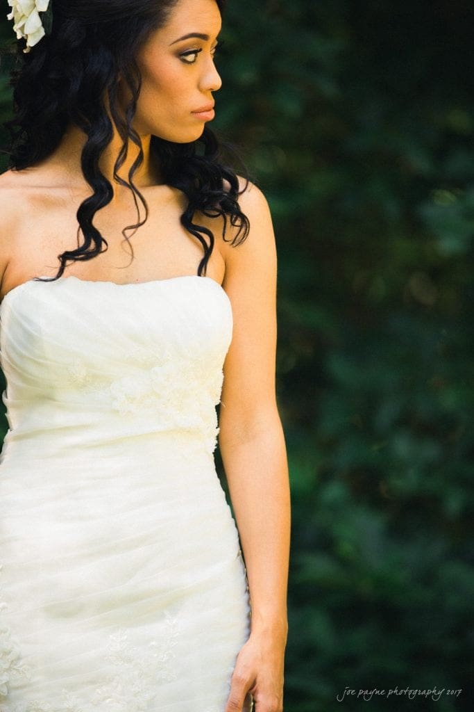 duke gardens wedding photography - jordan & zeke A bride in a white strapless gown stands outdoors, facing left. Her long, dark hair is styled in loose curls with a floral accessory. The background is a blurred mix of green foliage.