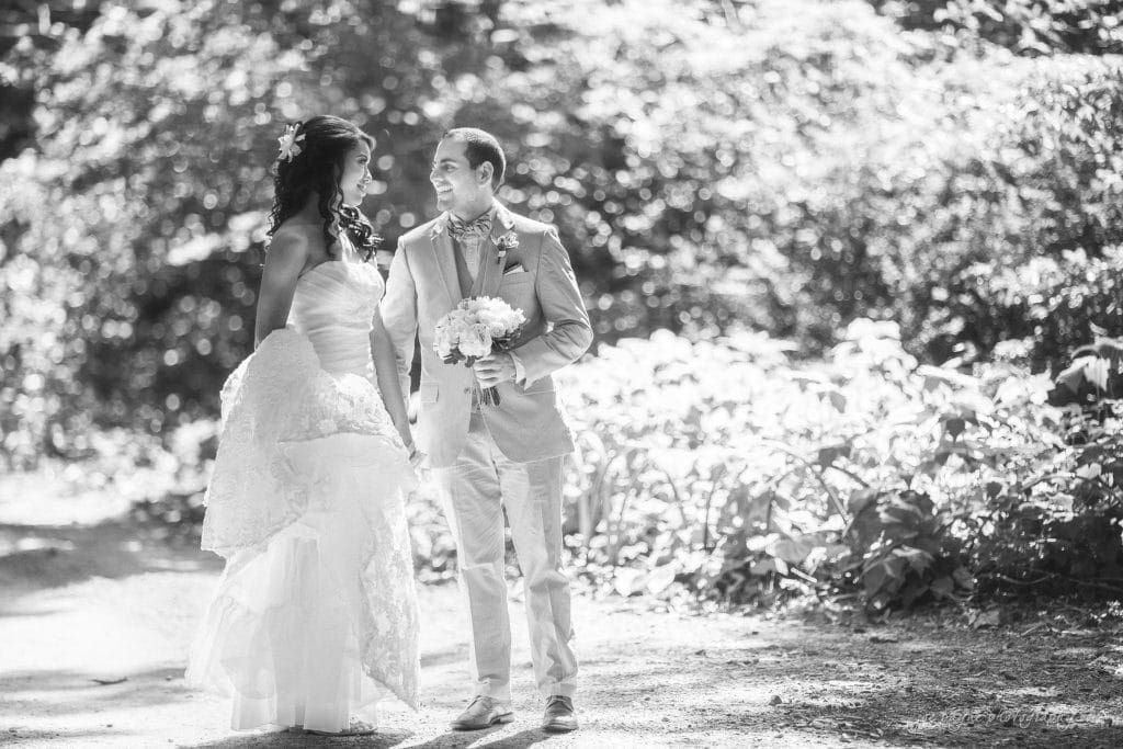 duke gardens wedding photography - jordan & zeke A bride and groom share a moment outdoors. The bride holds her lace gown and a bouquet, while the groom, in a light suit, smiles at her. They are surrounded by lush greenery in a sunlit setting, captured in black and white.