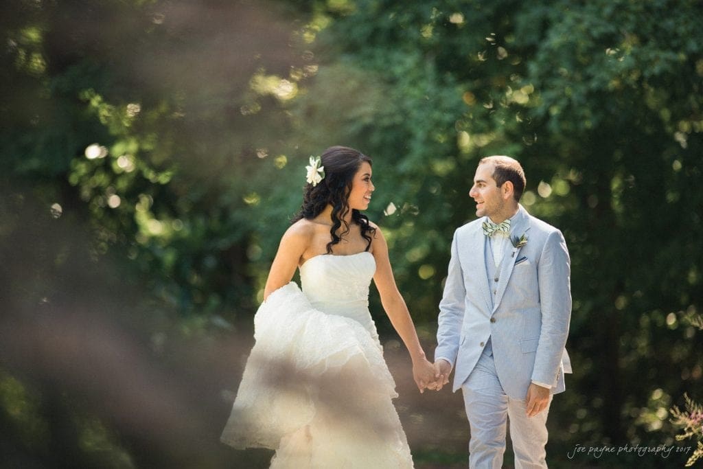 A bride in a white strapless gown and a groom in a light gray suit hold hands and smile at each other while walking outdoors, surrounded by lush green trees—perfect for Duke Gardens wedding photography.