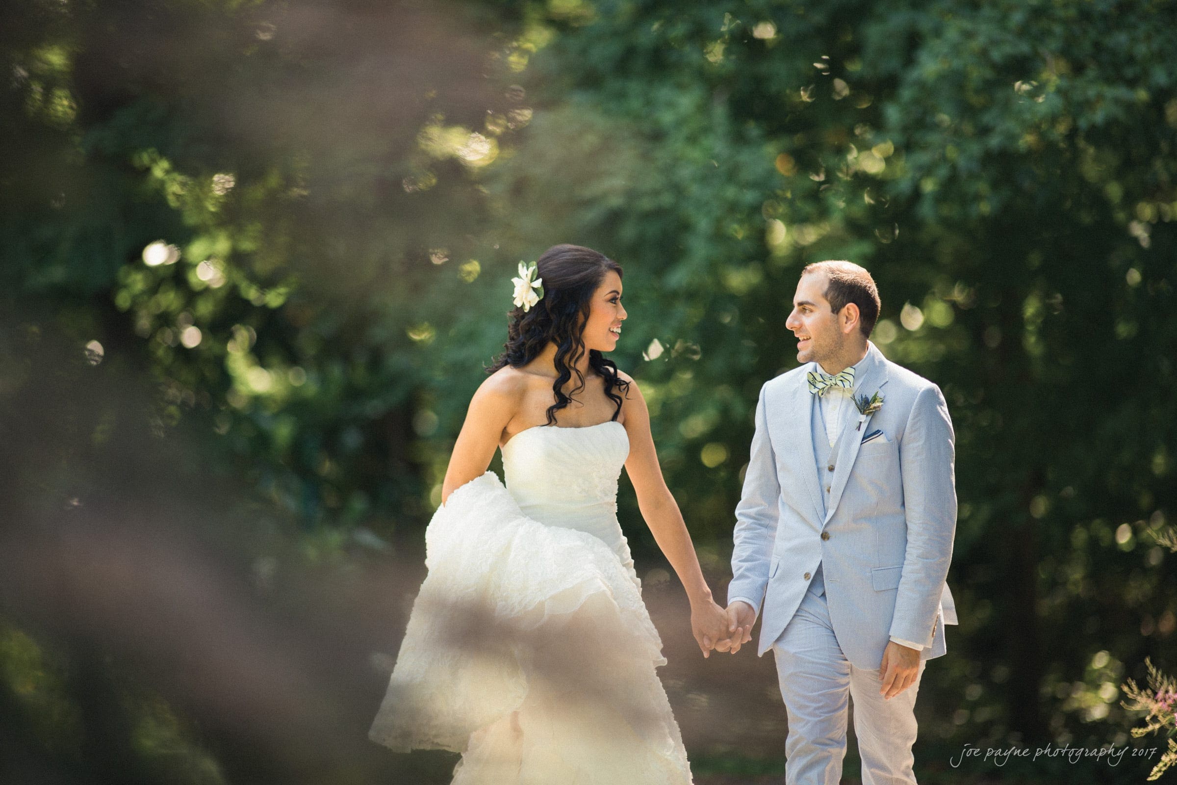 A bride in a white strapless gown and a groom in a light gray suit hold hands and smile at each other while walking outdoors, surrounded by lush green trees—perfect for Duke Gardens wedding photography.