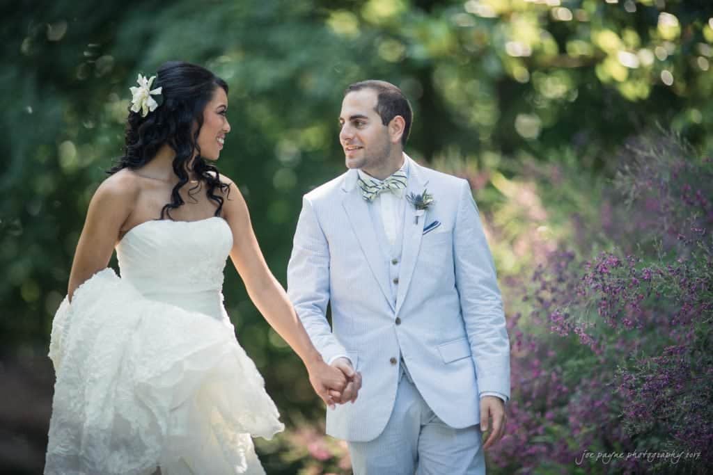 duke gardens wedding photography - jordan & zeke A bride in a flowing white gown and a groom in a light gray suit hold hands and smile at each other while walking through a garden. Lush greenery and blooming purple flowers surround them.