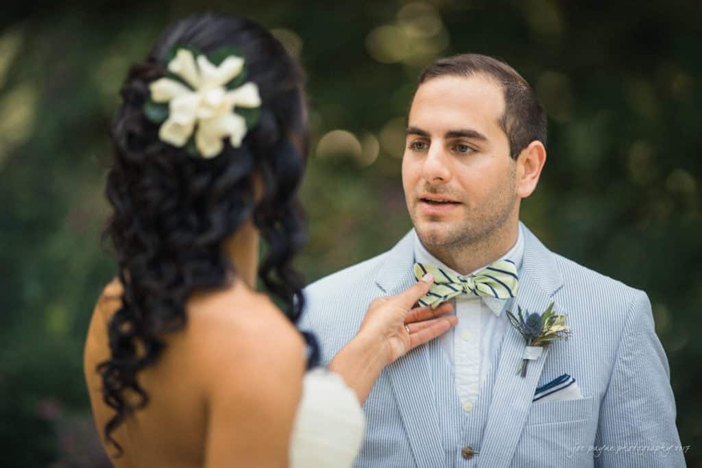 duke gardens wedding photography - jordan & zeke A groom wearing a light blue suit and striped bowtie gazes at a woman with long, dark, curly hair adorned with white flowers. The woman is adjusting his bowtie. The background is blurred greenery.