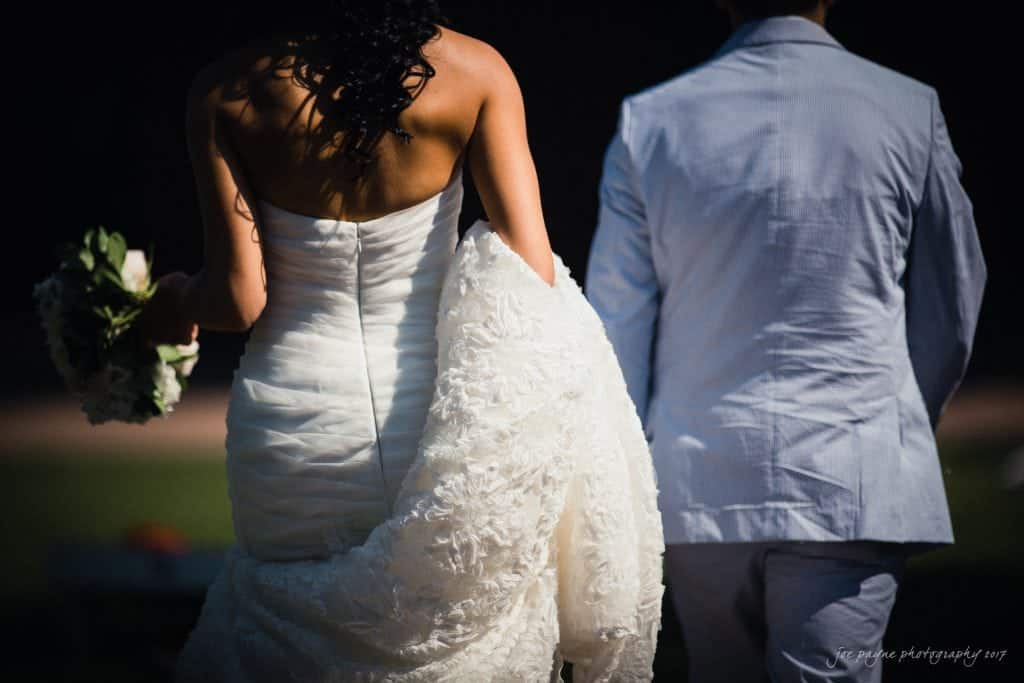 duke gardens wedding photography - jordan & zeke A bride in a strapless gown holding a bouquet and a groom in a light suit walk side by side, captured from behind. The scene is outdoor and sunlit, highlighting the dresss intricate texture.