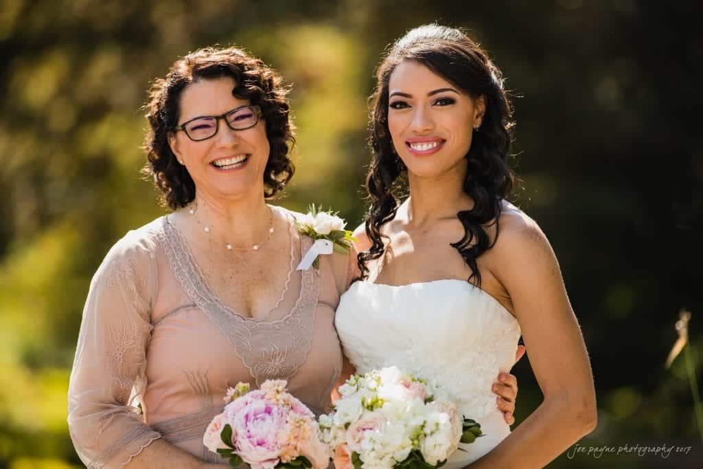 duke gardens wedding photography - jordan & zeke Two women smiling outdoors, wearing formal dresses and holding bouquets. The woman on the left wears glasses and a blush dress, while the woman on the right is in a white strapless gown. They stand against a backdrop of greenery.