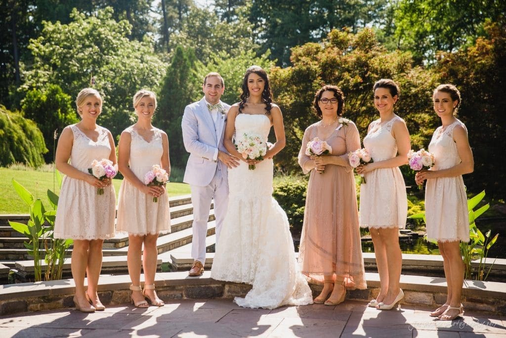 duke gardens wedding photography - jordan & zeke A bride and groom stand with five bridesmaids in a sunlit garden. The bride wears a white gown, and the groom is in a light suit. The bridesmaids wear light dresses and hold pink and white flowers. Trees and greenery are in the background.