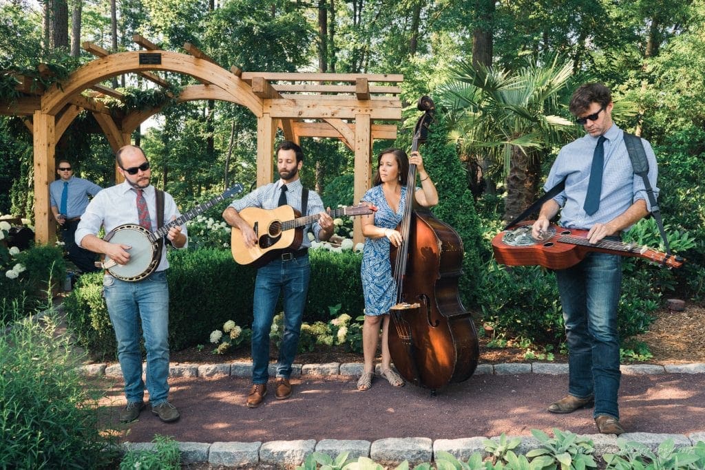 A bluegrass band performs in an outdoor garden setting with trees and wooden arches. The group includes a banjo player, two guitarists, and a bassist. Theyre casually dressed, and the sunlight filters through the lush greenery.