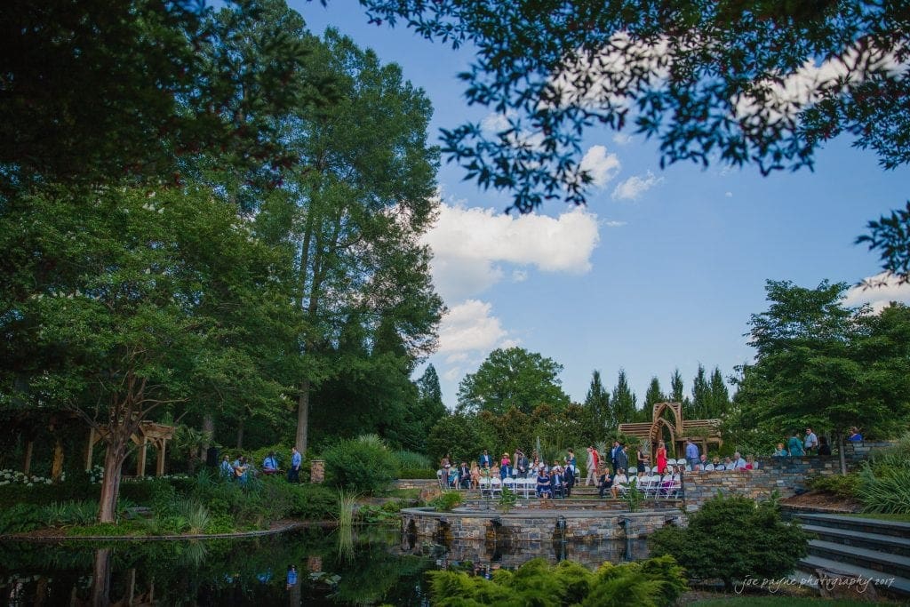 duke gardens wedding photography - jordan & zeke A scenic outdoor wedding ceremony set in a lush garden. Guests are seated on chairs arranged on a stone terrace, surrounded by tall trees and vibrant greenery. A small pond reflects the blue sky and scattered clouds overhead.