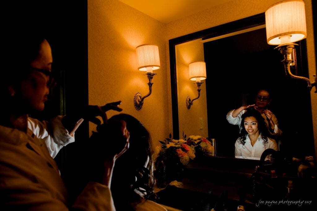 duke gardens wedding photography - jordan & zeke A woman smiles in a mirror as another person styles her hair in a warmly lit room. A bouquet of flowers sits on the counter beside them.