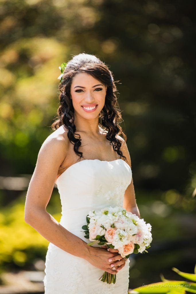 duke gardens wedding photography - jordan & zeke A bride in a white strapless wedding dress smiles while holding a bouquet of pink and white flowers. She stands outdoors with greenery in the blurred background, her dark hair cascading in curls.