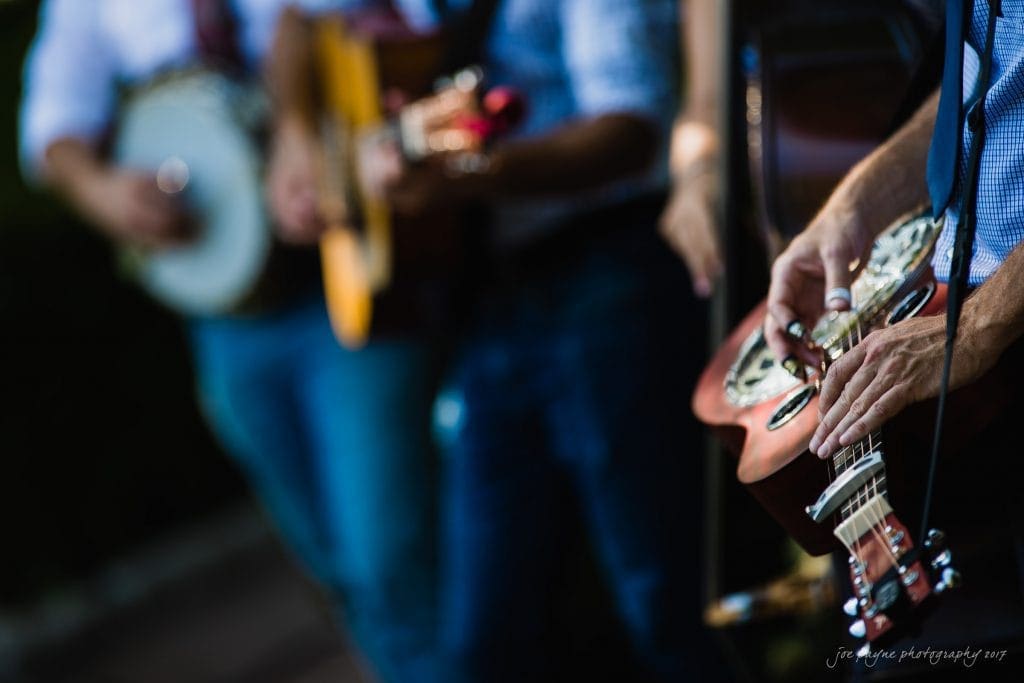 duke gardens wedding photography - jordan & zeke Close-up of musicians playing string instruments outdoors. Foreground shows hands on a resonator guitar with other guitarists and a banjo player visible in the background. Sunlight highlights the instruments against a blurred greenery backdrop.