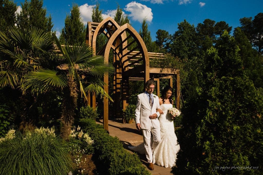 duke gardens wedding photography - jordan & zeke A bride and groom in elegant attire walk hand in hand down a garden path. The groom wears a white suit with a blue tie, and the bride holds a bouquet, wearing a white dress. They are surrounded by lush greenery and architectural structures.