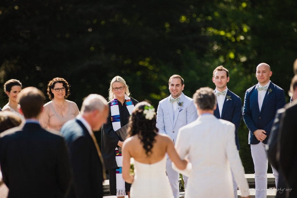 duke gardens wedding photography - jordan & zeke A wedding ceremony outdoors with a bride and groom facing each other, surrounded by a small group of smiling guests and a clergy member. Everyone is dressed formally, and the scene is set against a backdrop of greenery and sunlight.