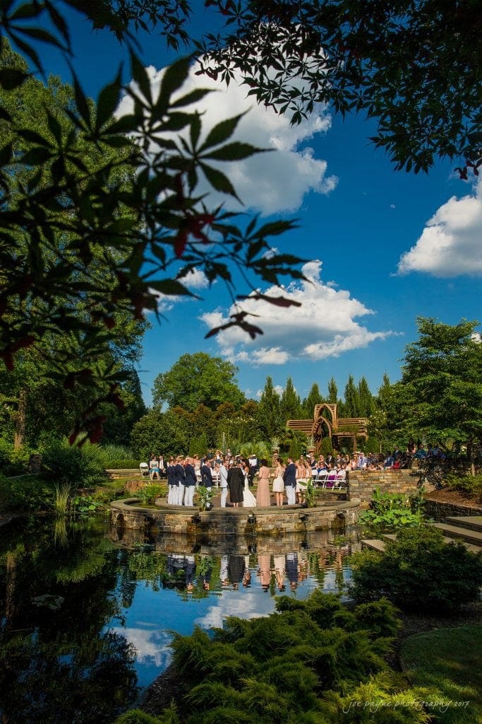 duke gardens wedding photography - jordan & zeke A scenic outdoor wedding ceremony takes place beside a pond, surrounded by lush greenery and under a bright blue sky with scattered clouds. Guests are seated around the couple, and the landscape reflects in the water.