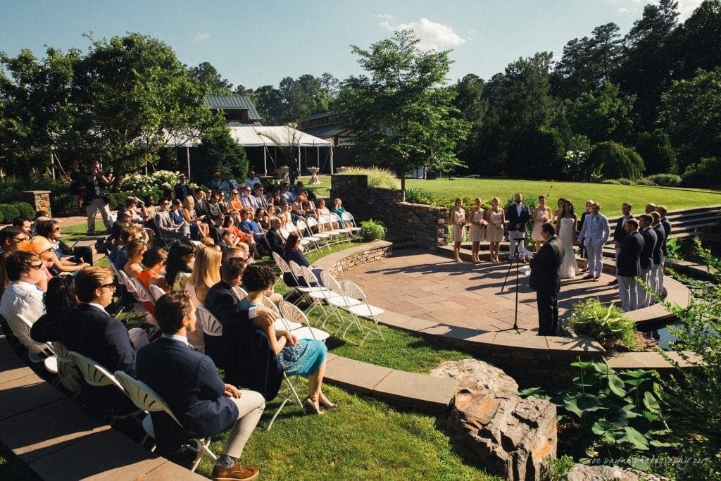 duke gardens wedding photography - jordan & zeke A wedding ceremony in a sunny outdoor setting with guests seated in white chairs arranged in an arc. The bridal party stands near the officiant at a small stone platform, surrounded by lush greenery and trees.